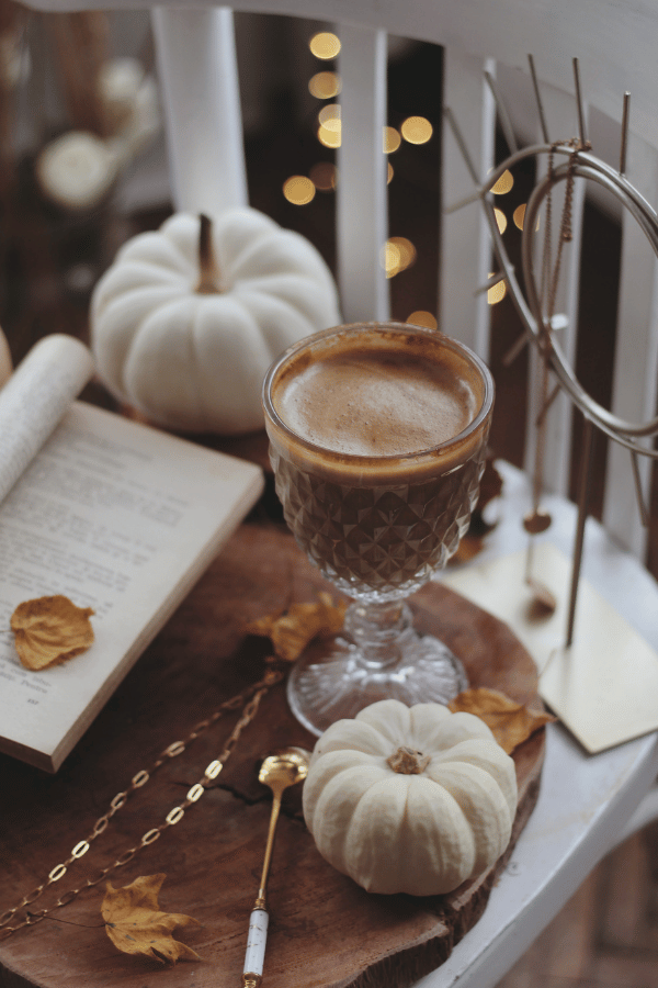 Warm cup of coffee on a wooden tray beside white pumpkins and a small open book, with scattered cinnamon sticks and a gold-speckled garland in the background