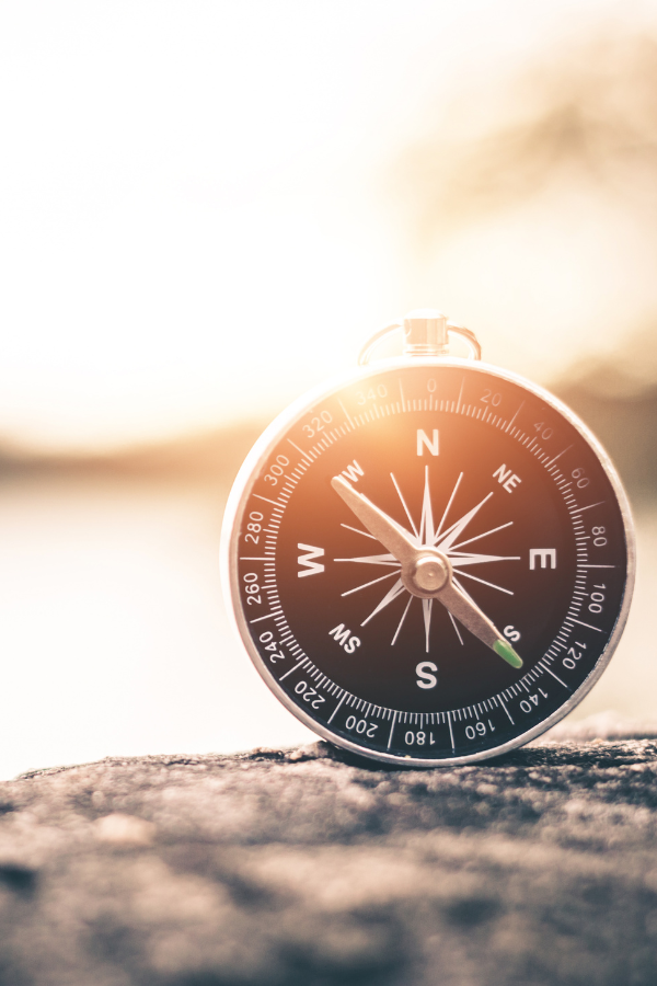 A close-up of a compass resting on a rocky surface with a blurred background of water and sky, illuminated by warm sunlight.