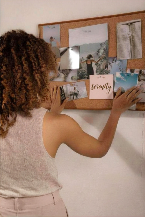 A person arranges photos, notes, and reflective materials on a cork vision board, including a card that reads “serenity.”