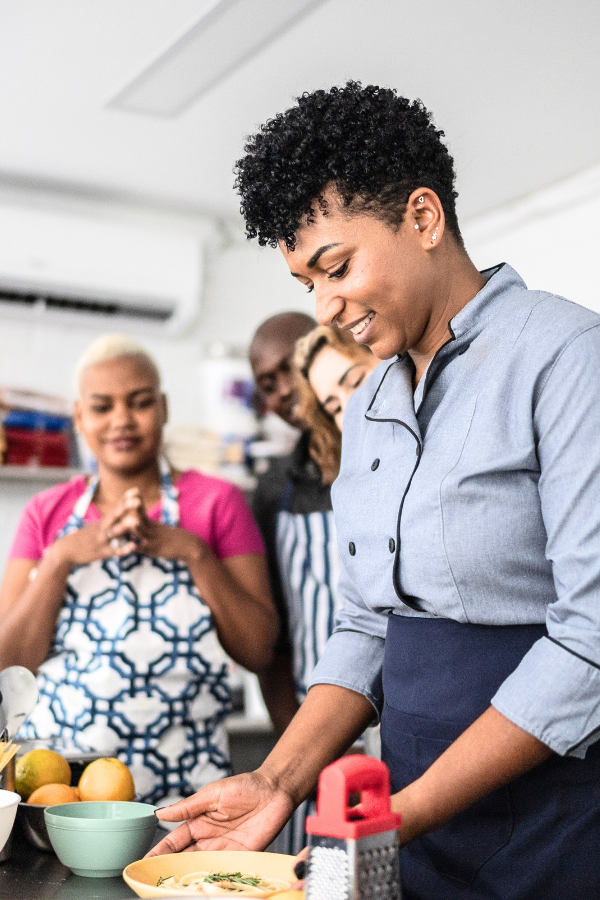 A small group in a bright kitchen watches as one person prepares food at a counter, with ingredients and tools arranged within reach.