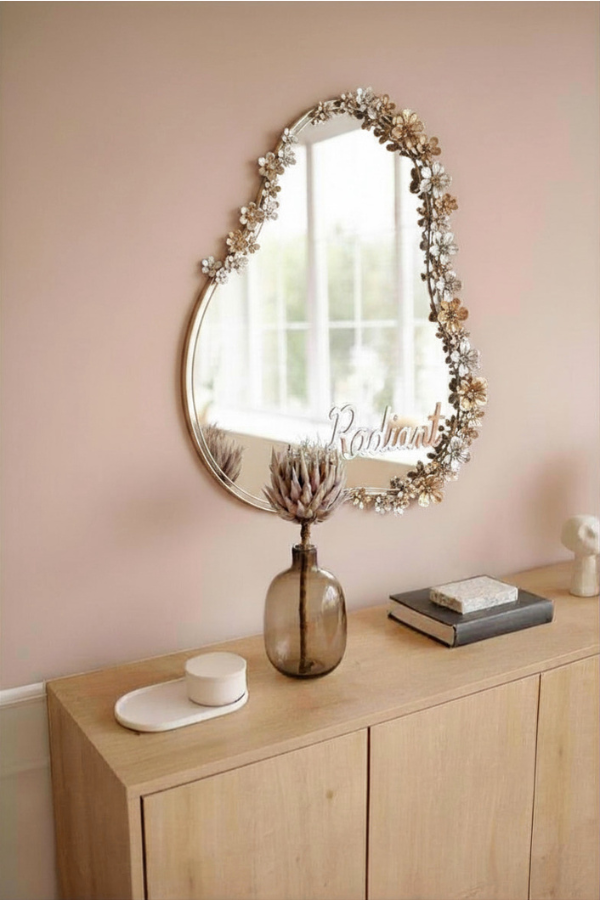 A decorative mirror with floral edges hangs above a wooden cabinet, reflecting a bright window. The word “Radiant” appears across the mirror.