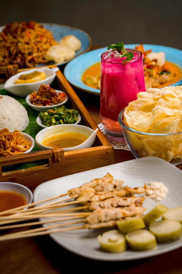 colorful spread of Indonesian dishes including grilled meat skewers, rice, small bowls of sauces and vegetables, crispy chips, and a bright pink drink arranged across a table.