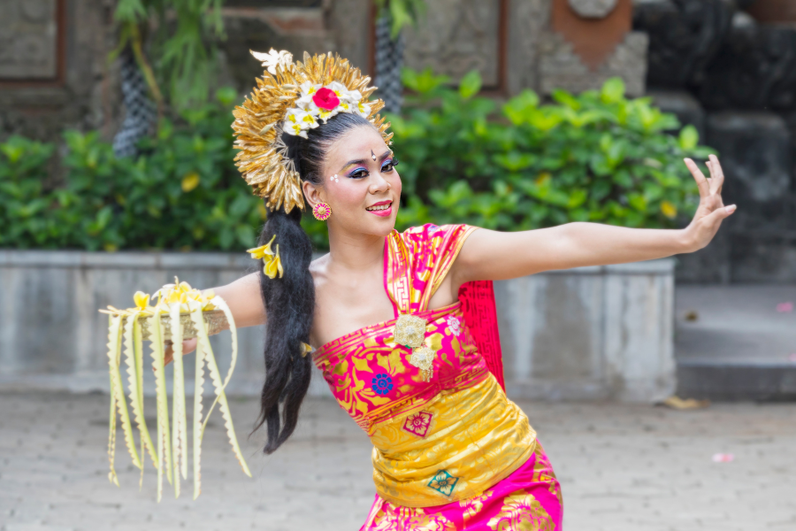 A Balinese Pendet dancer in vibrant orange and gold attire with an intricate headdress holds a bowl of flower petals, her hands poised in an expressive gesture.