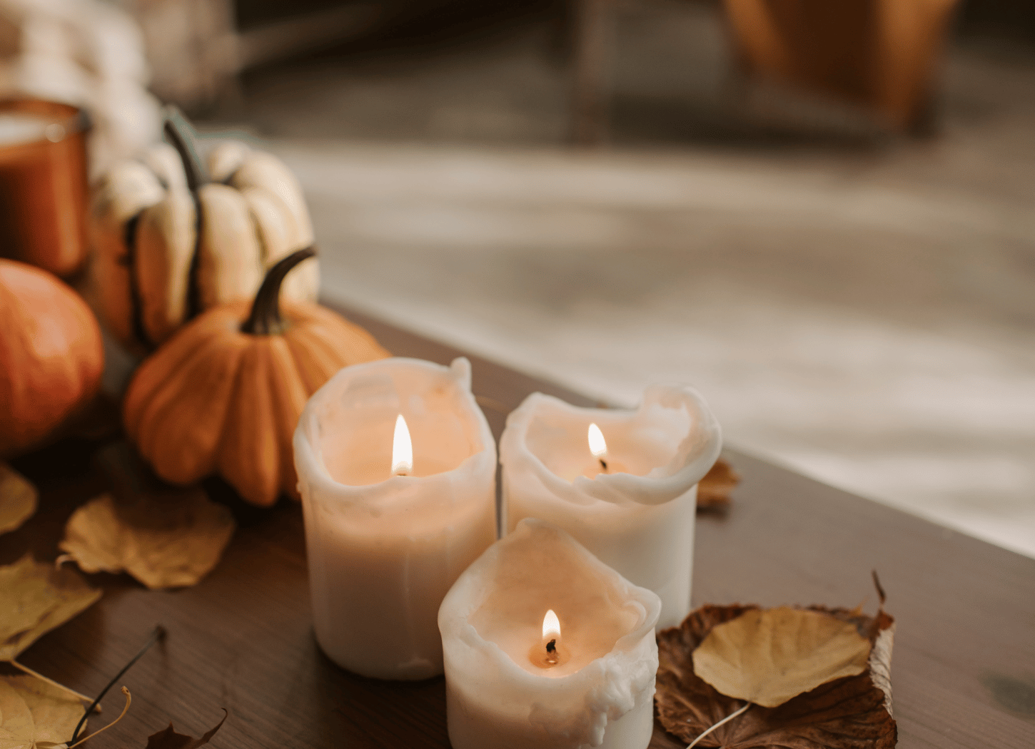 Three small white candles, scattered autumn leaves, and pumpkins on a wooden table and a chair in the background