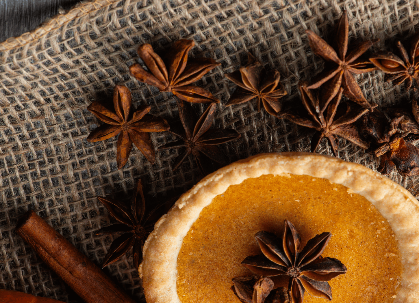A small round tart on burlap fabric, topped with a star anise spice, surrounded by scattered star anise pods and a cinnamon stick