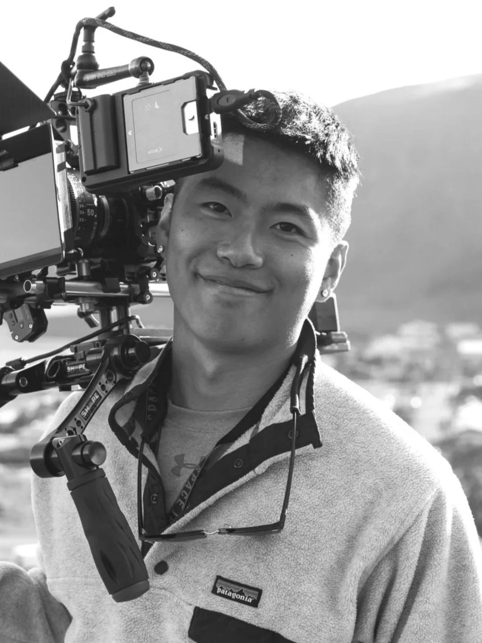 A young man with short hair holding a professional video camera on a shoulder rig outdoors, smiling, with a rural landscape and hills in the background.