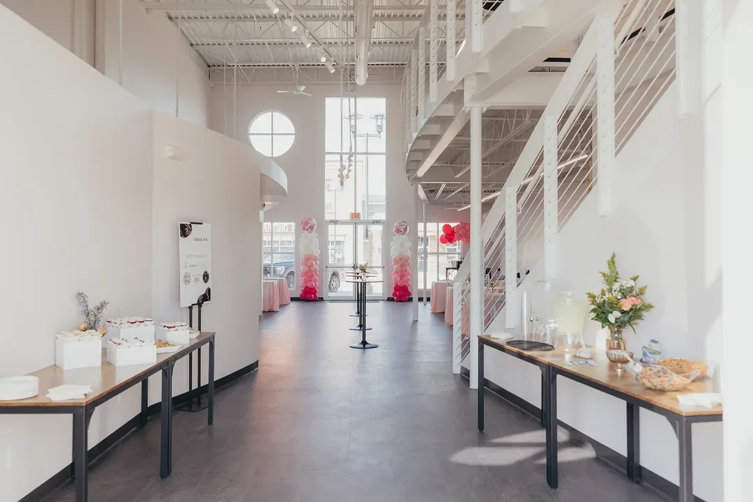 Interior of a modern event space with white walls, high ceilings, and large windows. There are decorated tables with pink and red balloons near the entrance, and a refreshment table with flowers and snacks on the right side.