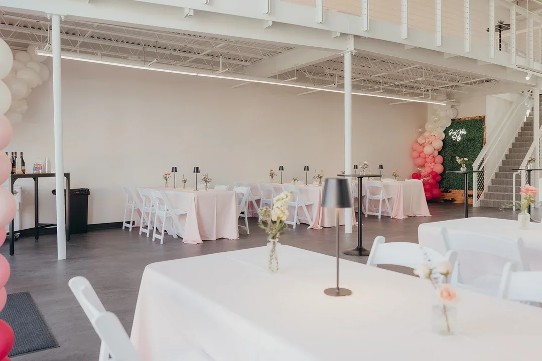 Event space decorated for a celebration with white tables, pink tablecloths, flower centerpieces, and balloon decorations in pink and white.