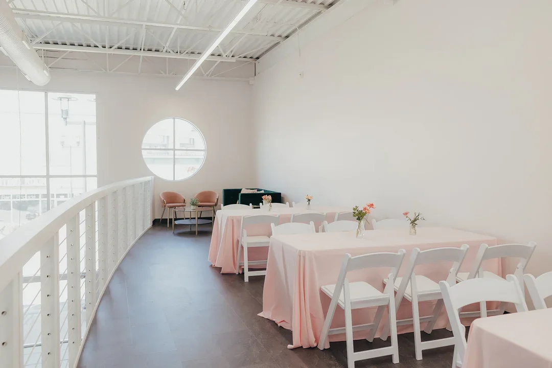 A bright, modern event space with white walls, pink tablecloths, and white chairs. Small vases with flowers are on the tables, and there are pink and black chairs against the wall near a circular window. Large windows let in natural light.