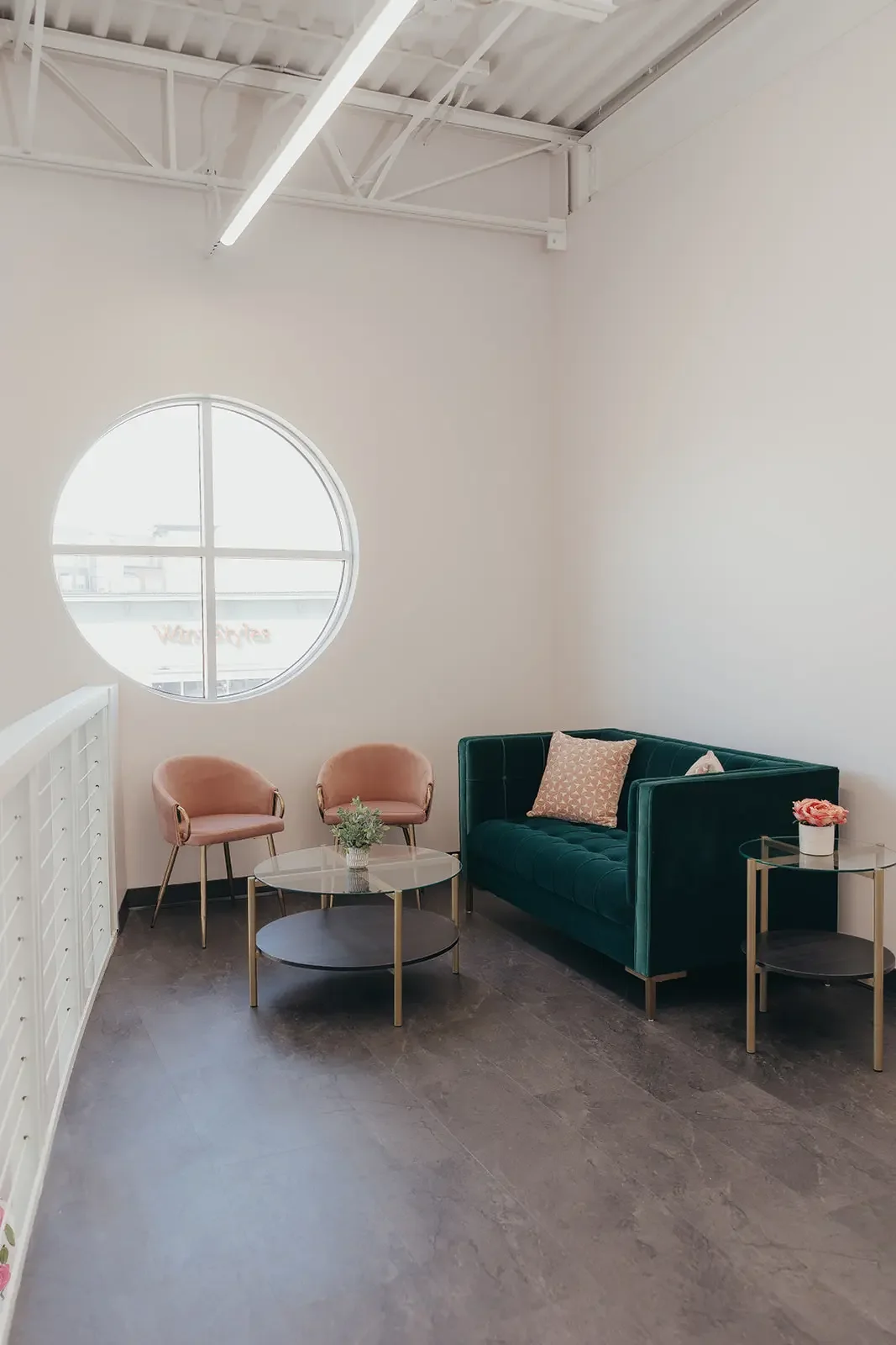 Modern waiting area with a green velvet sofa, two blush pink armchairs, two glass and metal side tables, and a round window on a white wall.