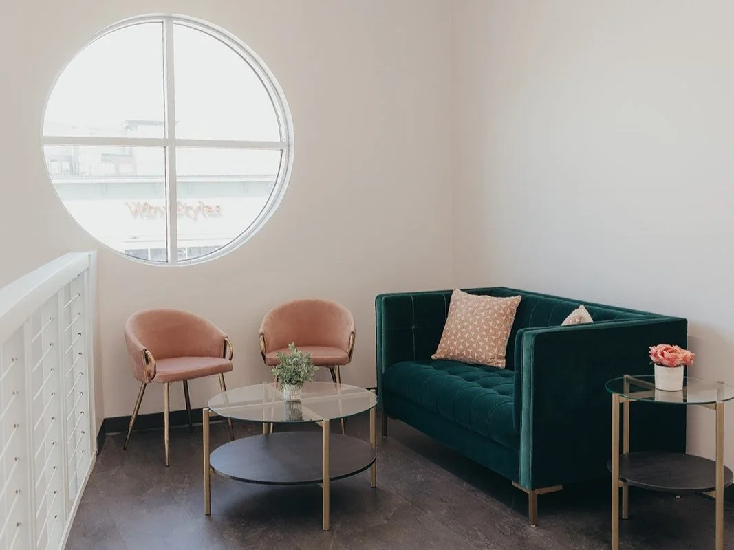 A cozy sitting area with a dark green velvet couch, two pink armchairs, a round coffee table, and side tables with flowers, near a large round window letting in natural light.