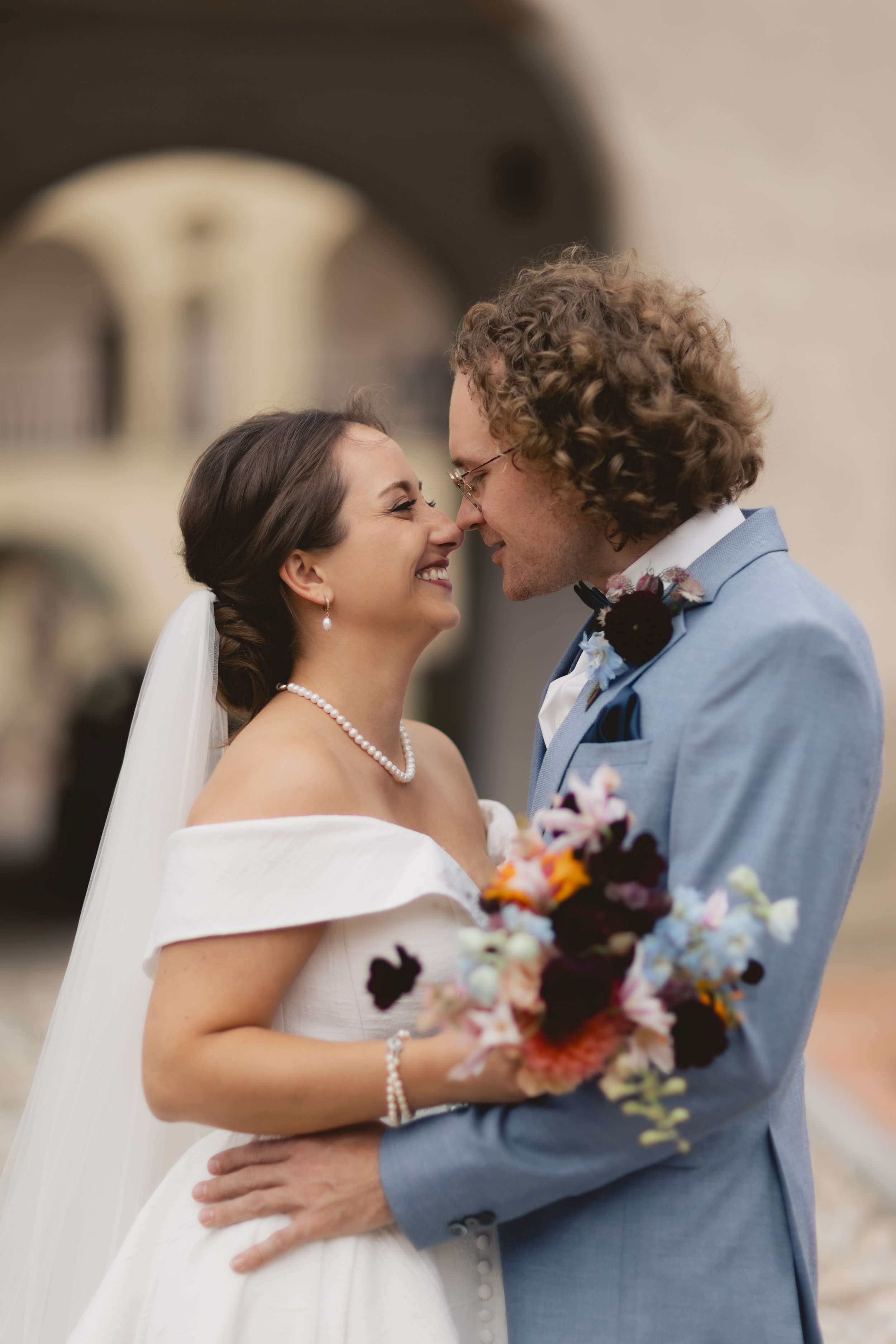 Ein Hochzeitsbild eines Paares, das sich anlächelt und bei ihrer Trauung eng umarmt, mit starkem emotionalem Ausdruck, im Freien, im Hintergrund eine historische Gebäude-Architektur.