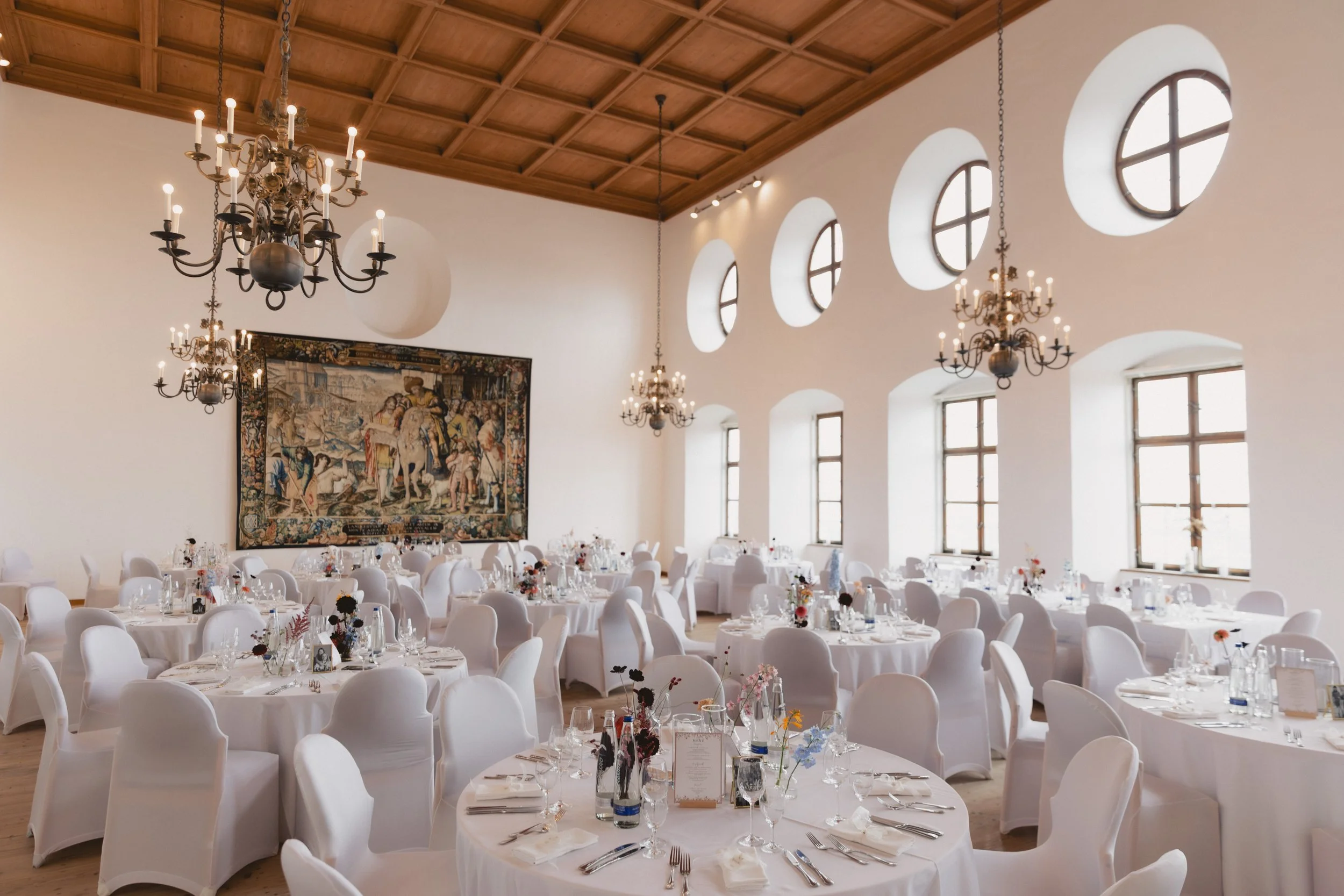 Eleganter Veranstaltungsraum mit weißen Tischen und Stühlen, Kronleuchtern, großen Fenstern und Wandteppich.