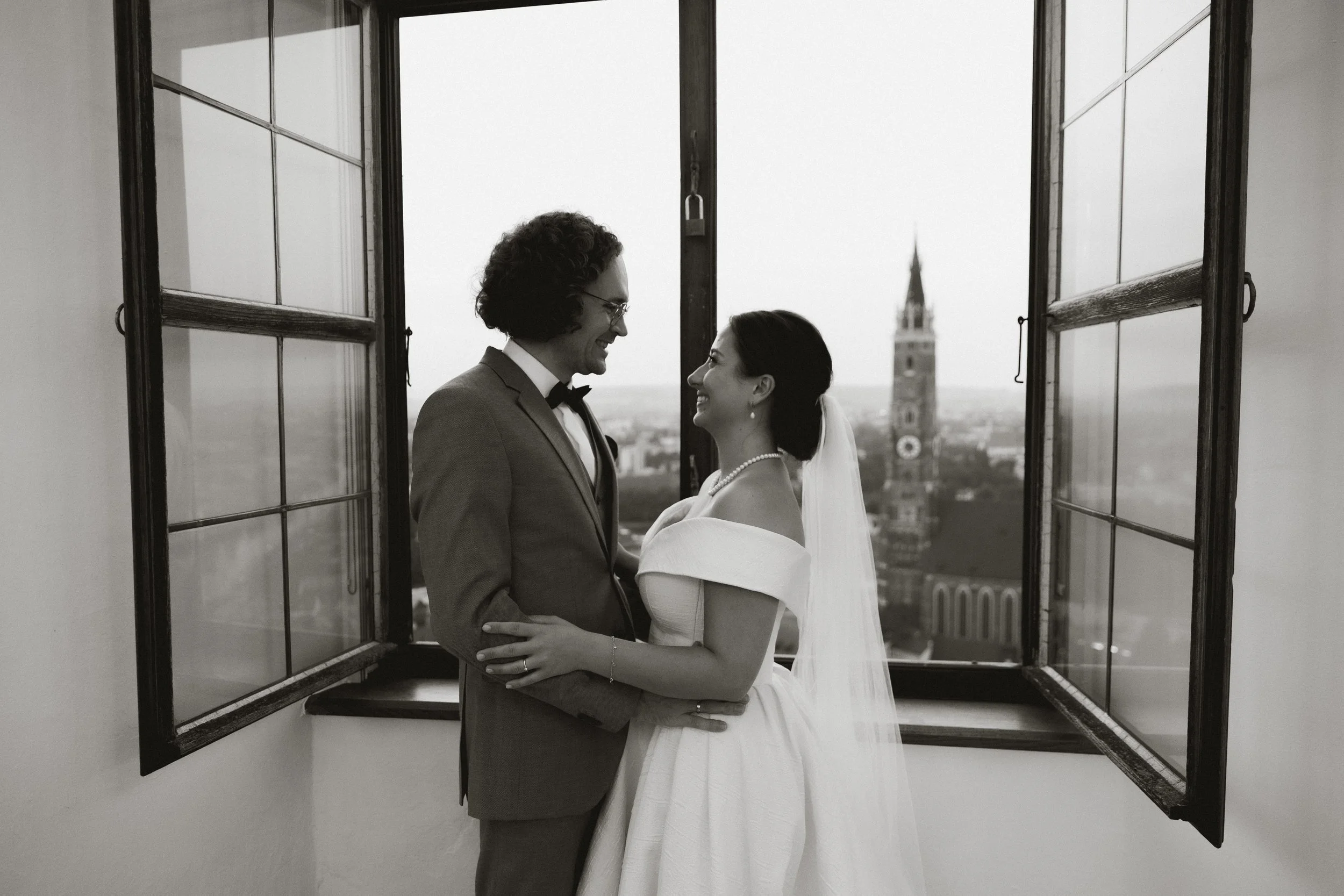 Ein Brautpaar steht vor einem Fenster und blickt sich lächelnd an, im Hintergrund die Stadtsilhouette mit einem Kirchturm.