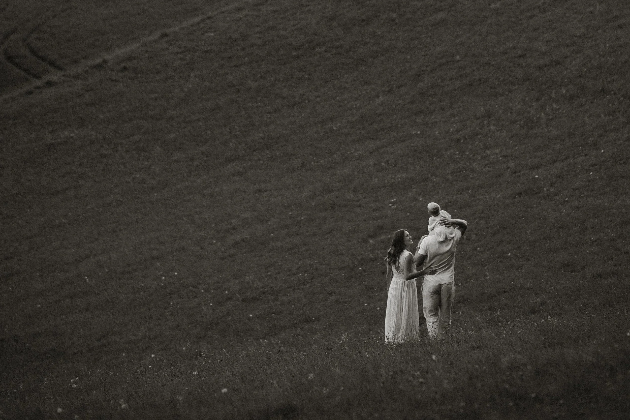 Eine Familie besteht aus einem Mann, einer Frau und einem Baby. Sie stehen zusammen in einer offenen, leicht ansteigenden Wiese bei Sonnenuntergang, in Schwarz-Weiß aufgenommen.