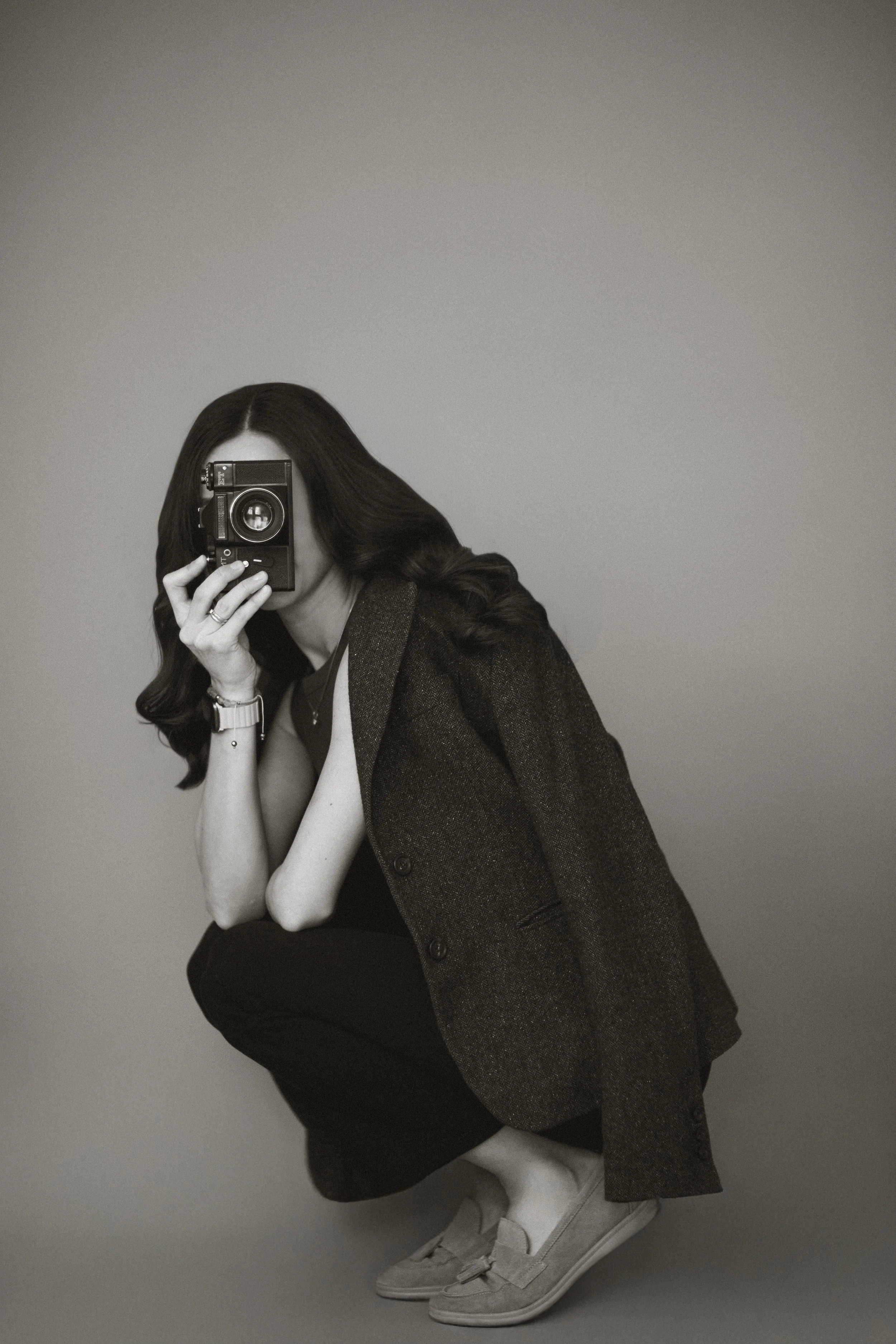 Schwarz-weiß-Foto einer Frau mit langen, dunklen Haaren, die in die Kamera eines alten Films zu fotografieren scheint, während sie in einer Squat-Position ist und eine Jacke trägt.