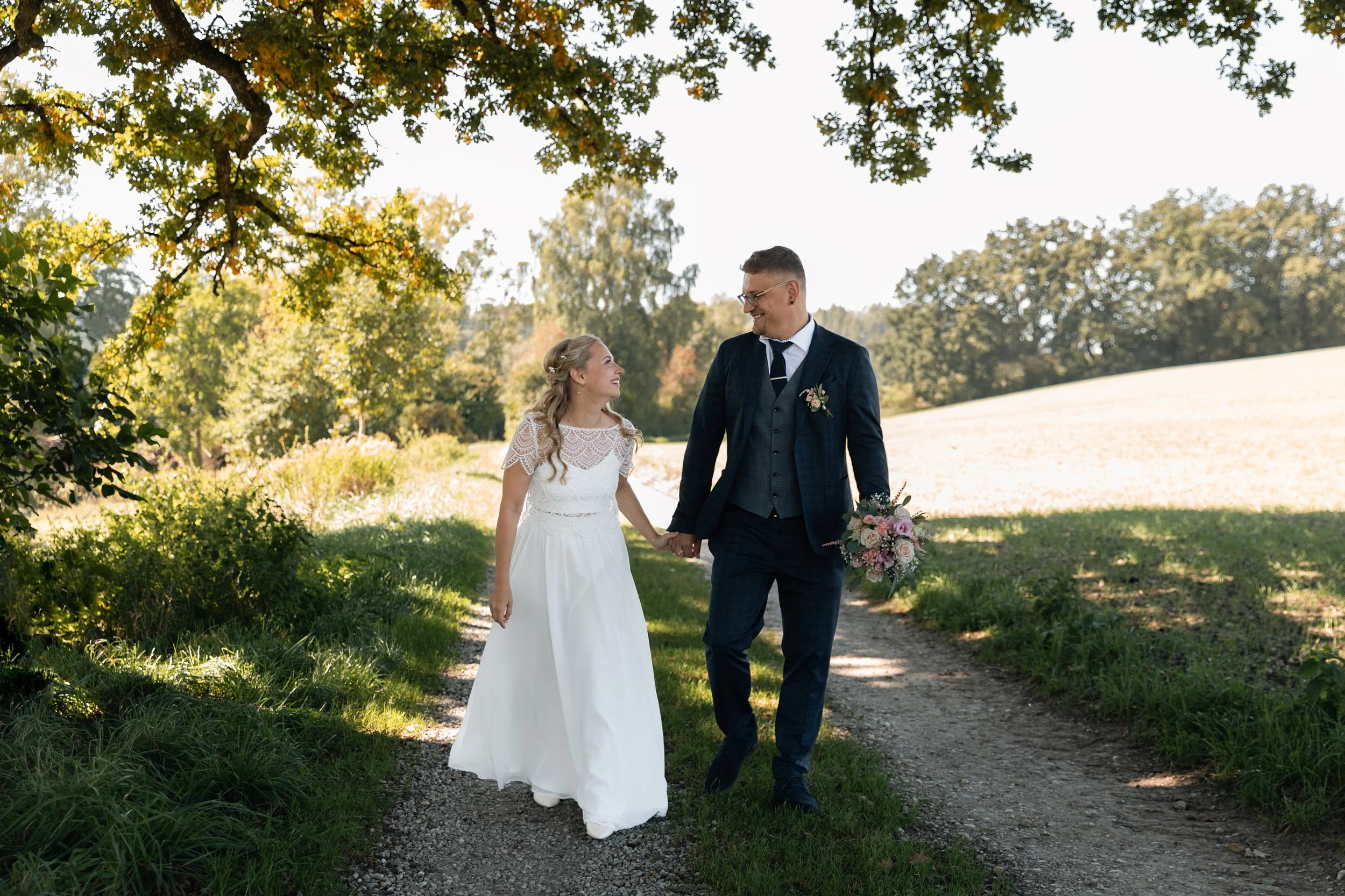 Ein verheiratetes Paar in Hochzeitskleidung läuft Hand in Hand auf einem Waldweg im Freien, umgeben von Bäumen und Natur, die Sonnenstrahlen scheinen durch die Blätter.
