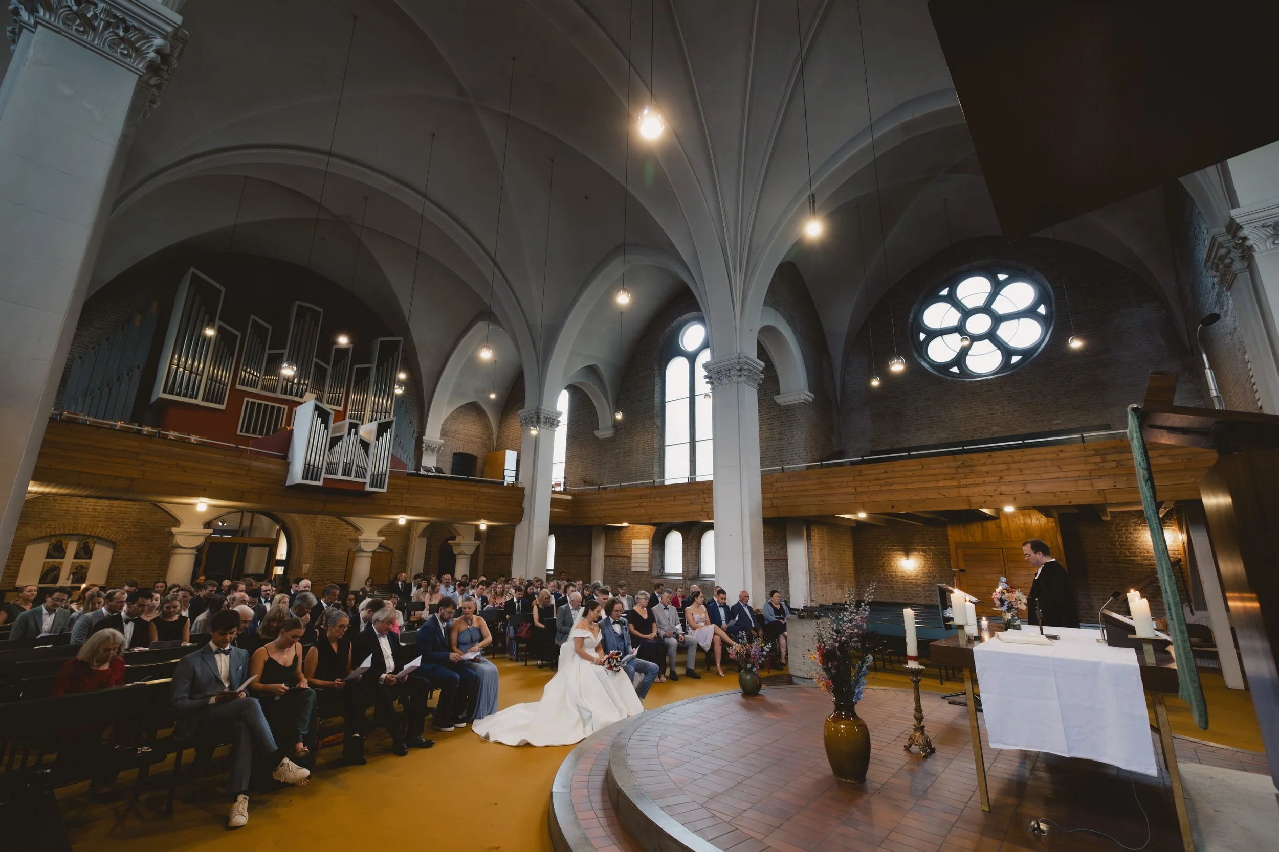 Hochzeit in einer Kirchenhalle mit Gästen, Braut in weißem Kleid, Pfarrer liest während der Trauung. Viele Kerzen, Vasen mit Blumen, Orgel im Hintergrund.