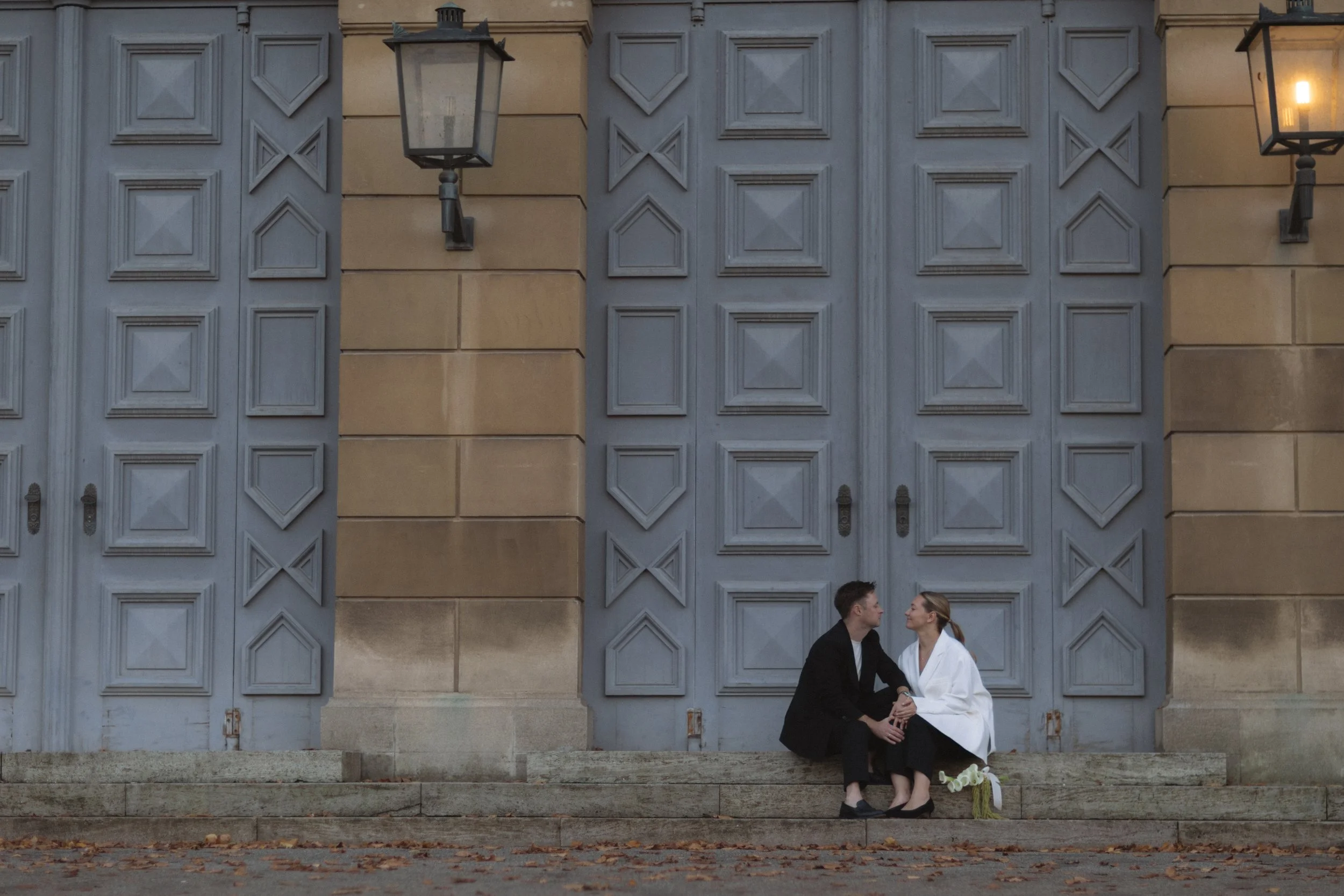Ein Mann und eine Frau sitzen vor einem großen blauen Tor, scheinen sich zu lieben, die Frau trägt ein weißes Kleid, der Mann einen schwarzen Anzug, umgeben von Laternen und Herbstblättern.