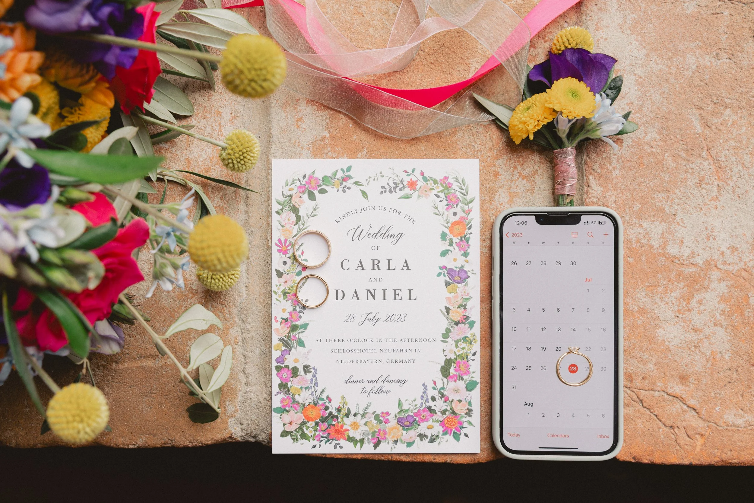 Ein Einladungskarte zur Hochzeit, zwei goldene Eheringe, ein Smartphone mit Kalenders App, bunte Blumen und eine Schleife auf einem braunen Steinuntergrund.