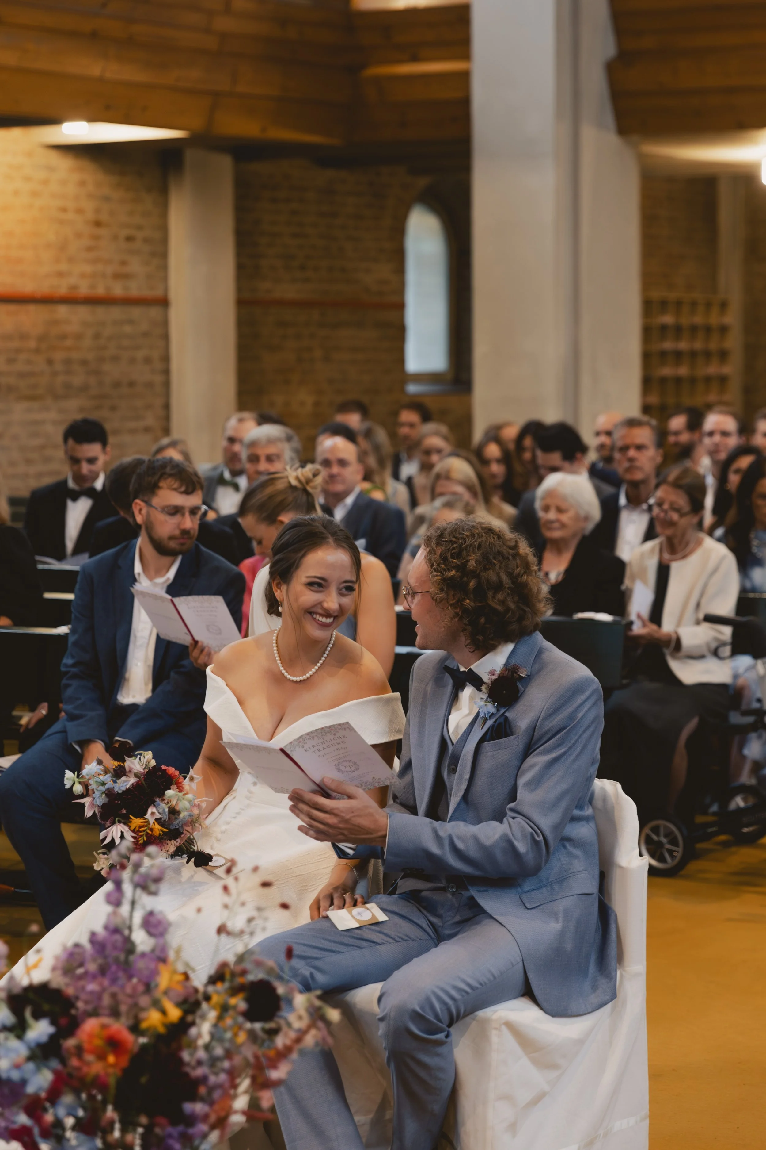 Hochzeitspaar sitzt im Vordergrund eines Saals, der mit Gästen gefüllt ist, und liest Trauungsurkunden, die sie anlächelt.