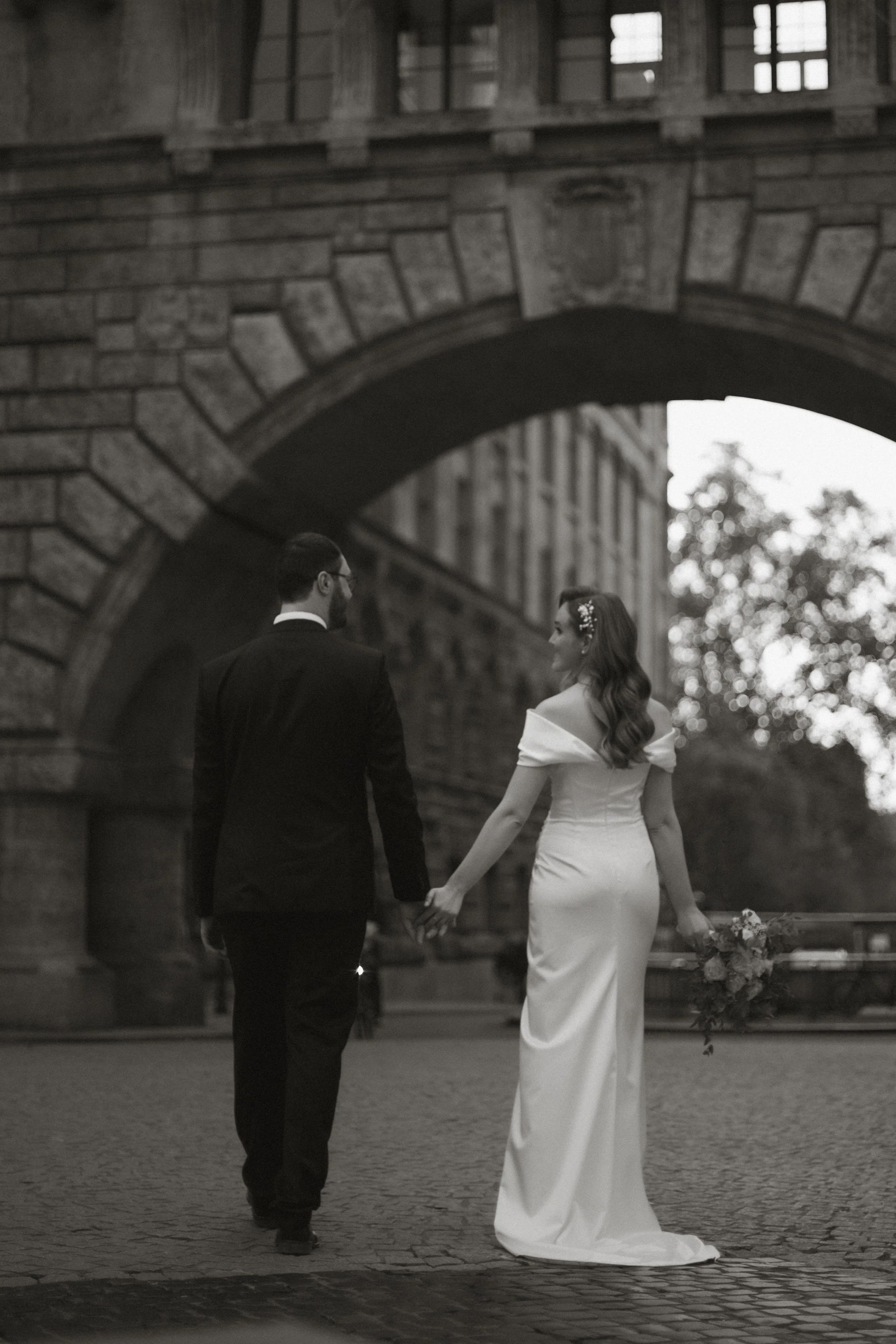 Ein Brautpaar in formeller Kleidung hält Händchen vor einer Steinbrücke.
