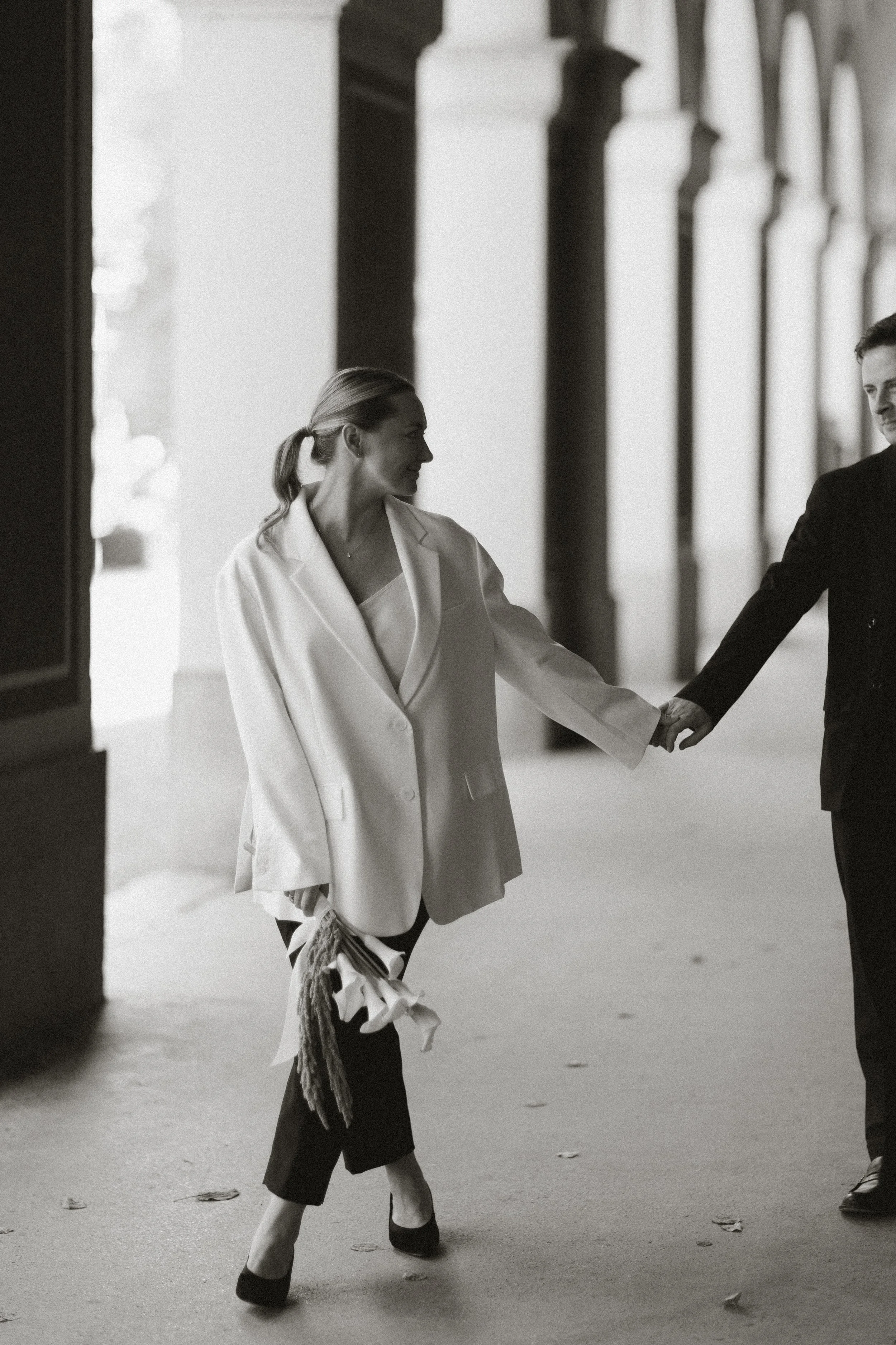 Zwei Menschen, eine Frau und ein Mann, halten sich an den Händen und gehen durch einen Bogen in einer Architektur mit Säulen, surreale Schatten und Lichteffekte, schwarz-weißer Stil.