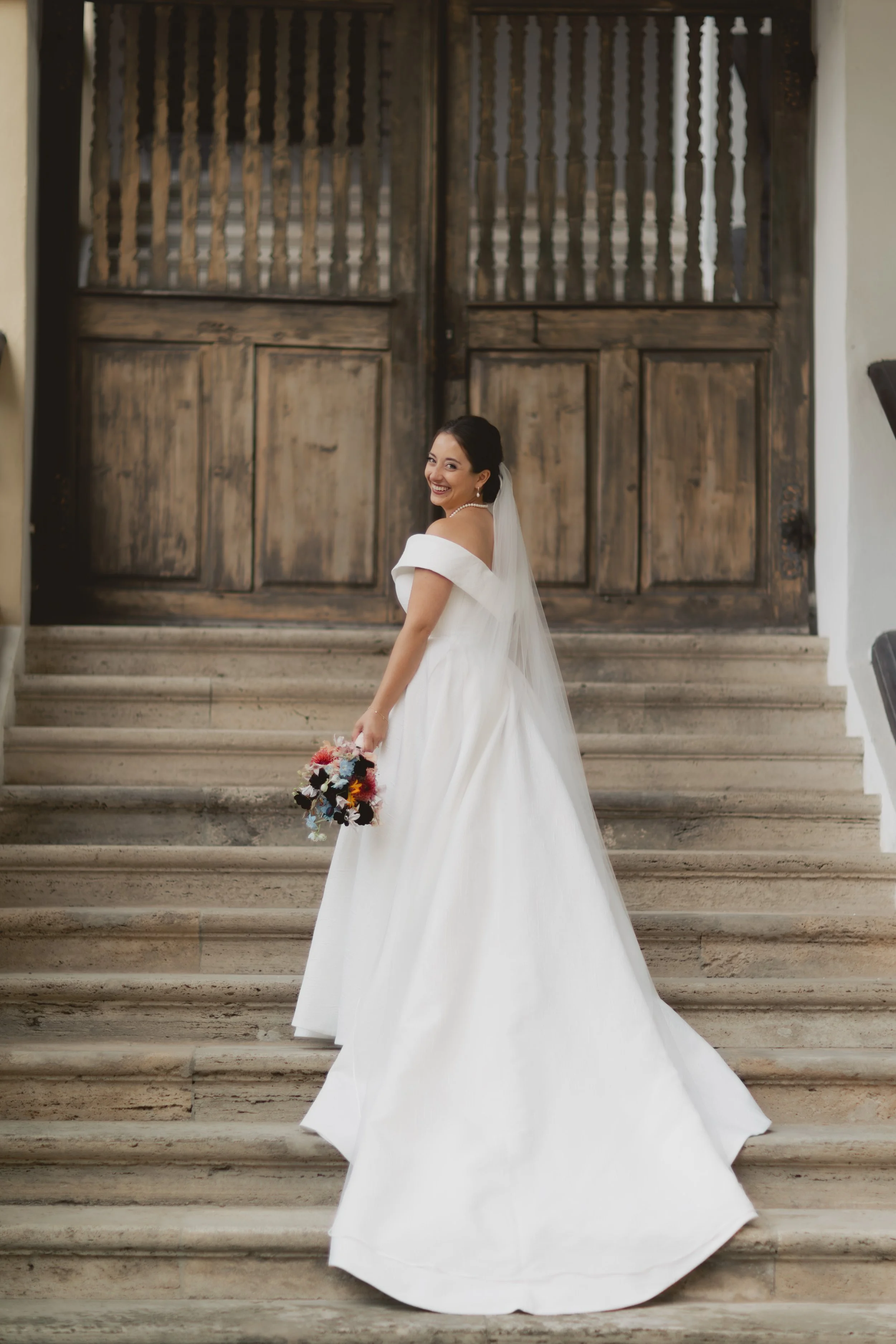 Braut in Weißes Brautkleid mit Blumenstrauß auf Treppe vor alten Holz-Tor.