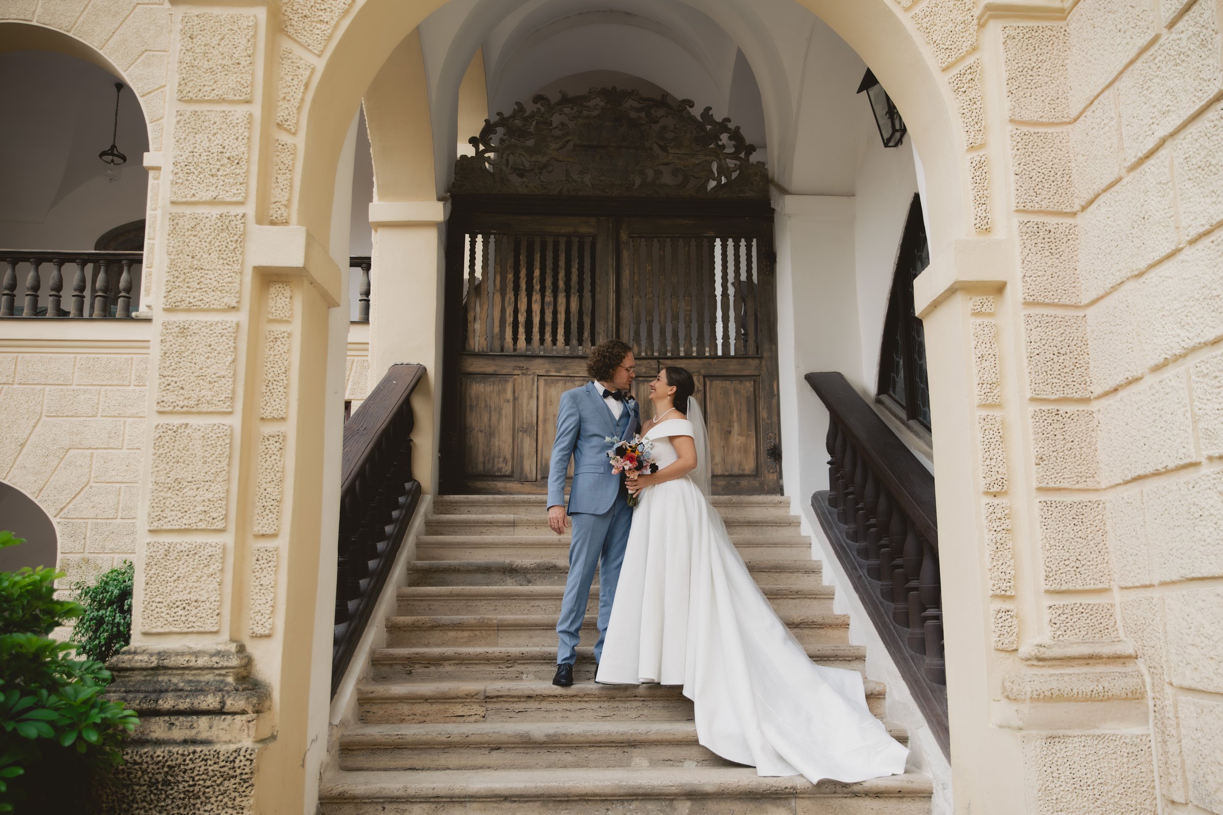 Ein Brautpaar steht auf Stiegen vor einer historischen Tür, das Brautkleid ist lang und weiß, der Bräutigam trägt einen blauen Anzug mit Fliege, beide blicken sich liebevoll an.