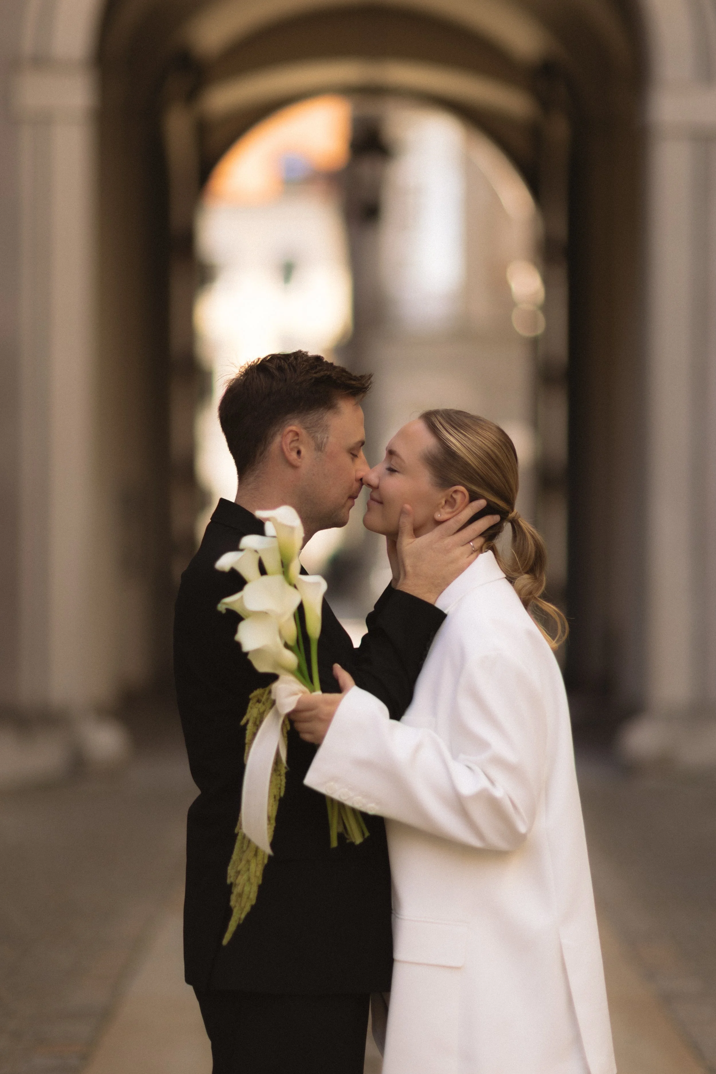 Ein Paar steht sich in einer Bogenpassage gegenüber, sie hält einen Blumenstrauß, sie küssen sich zärtlich.