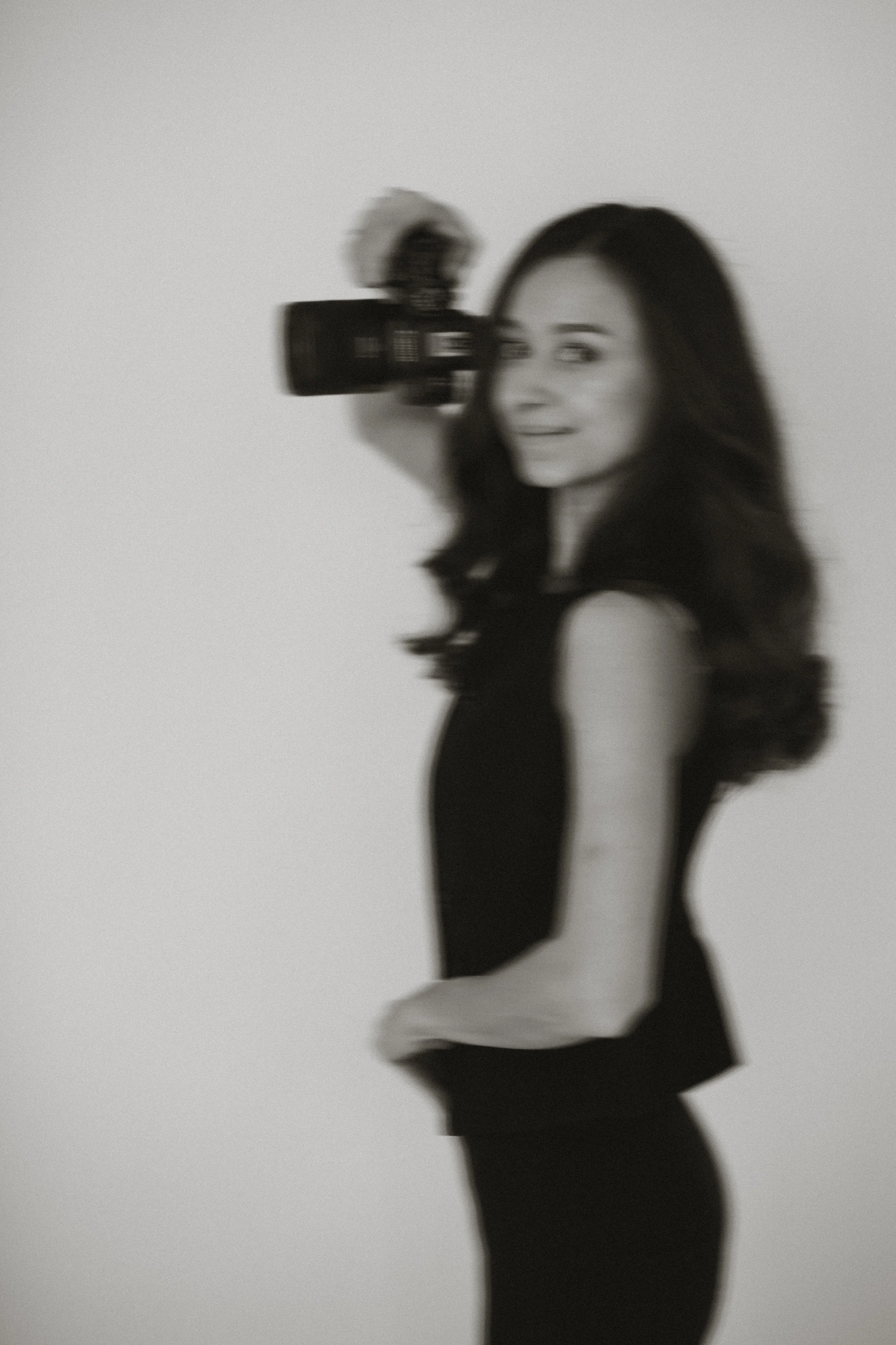 Schwarz-weiß Bild einer lachenden Frau mit dunklen, lockigen Haaren, die eine Videokamera hält, während sie in die Kamera schaut.