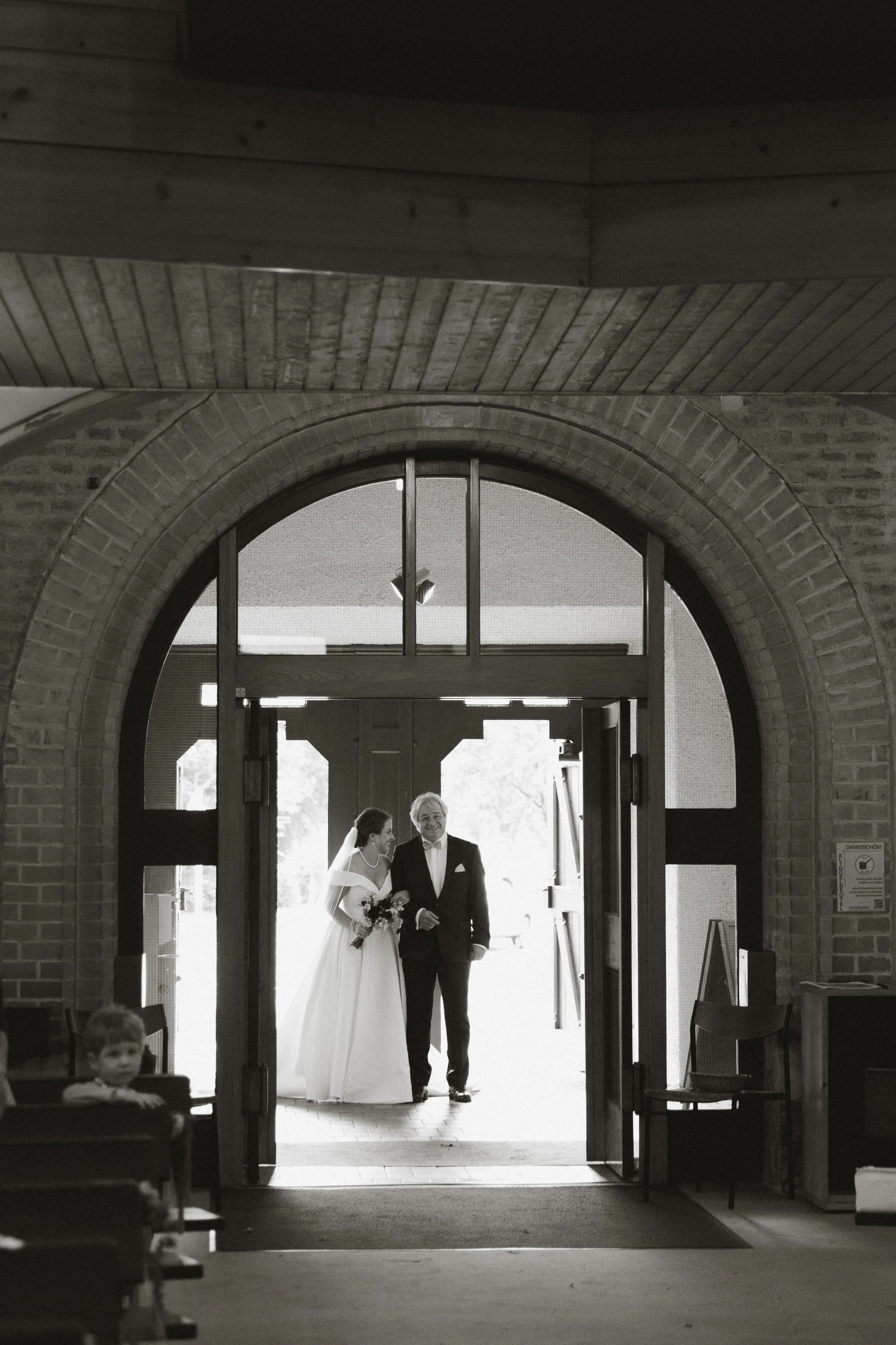 Braut in Hochzeitskleid und Bräutigam in Anzug beim Verlassen der Kirche, im Hintergrund geöffnetes Kirchentür, im Vordergrund kleiner Junge auf Bänken.