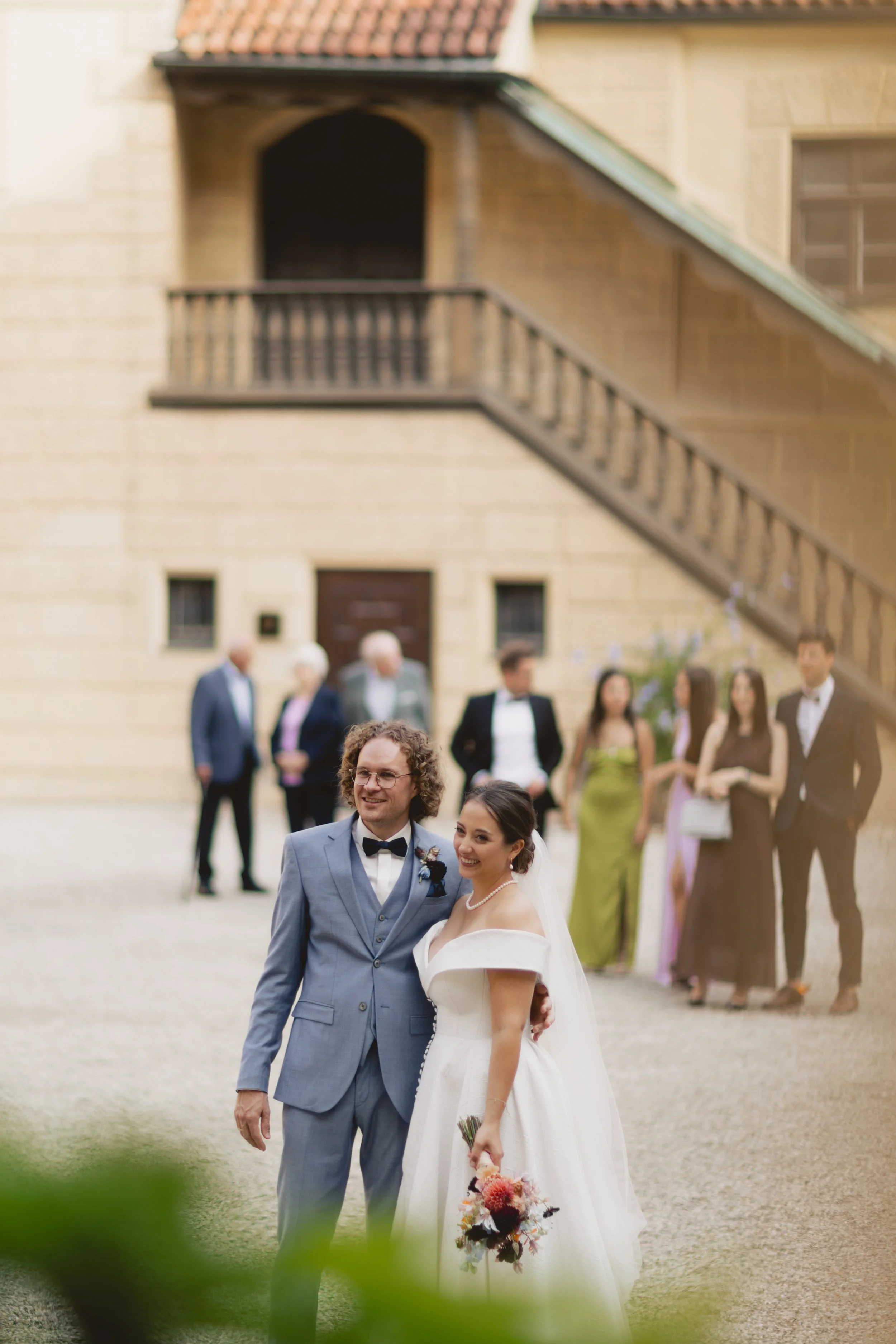 Braut und Bräutigam stehen zusammen in einem Brautkleid und einem Anzug, umgeben von Freunden und Familie bei einer Hochzeit im Freien vor einem alten Haus.