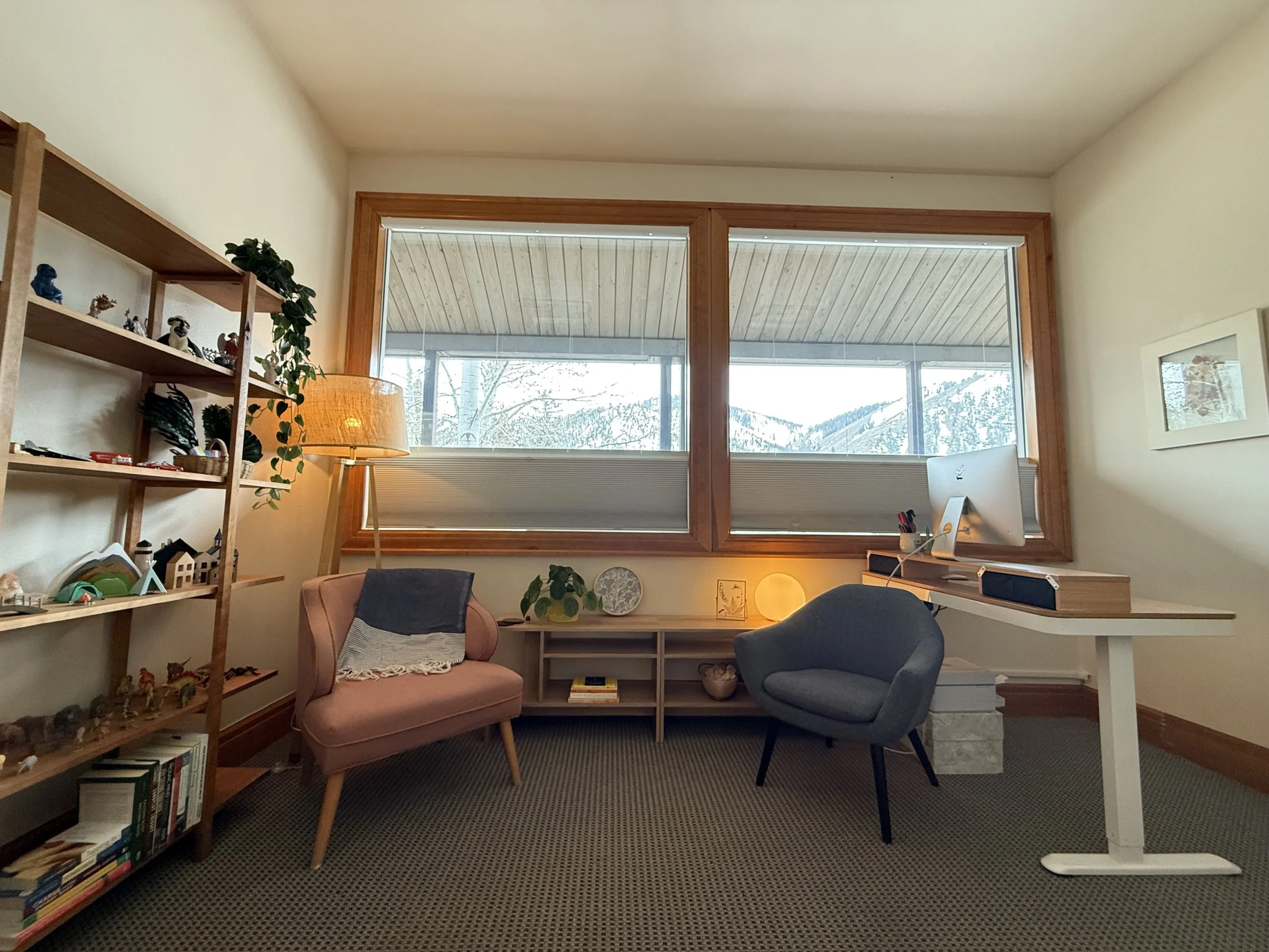 A cozy home office with a large window showing a snowy landscape outside. The room has a pink armchair, a grey chair, a wooden desk with a Mac computer, and shelves with decorative objects and books.
