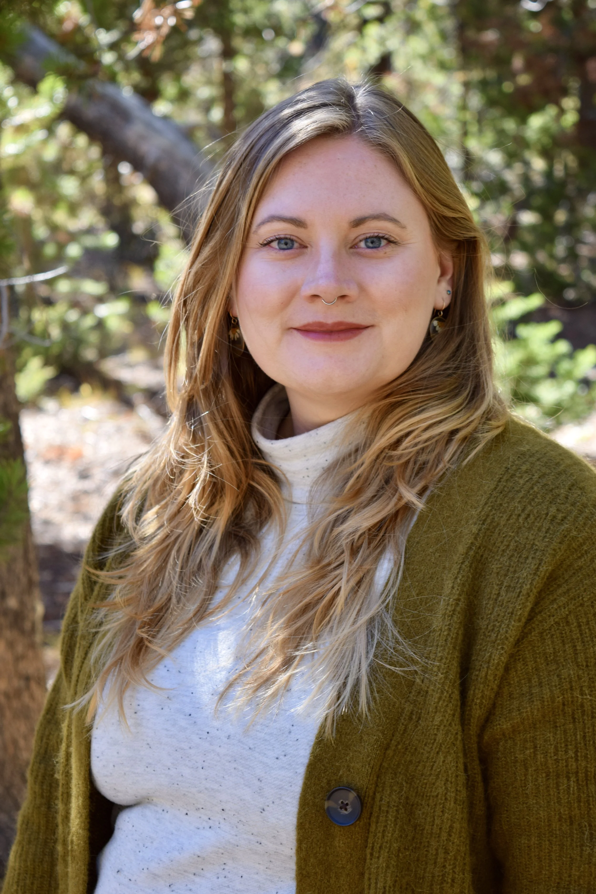 Close-up of a young woman with long, wavy blonde hair, blue eyes, and a septum piercing, standing outdoors with trees and sunlight in the background.