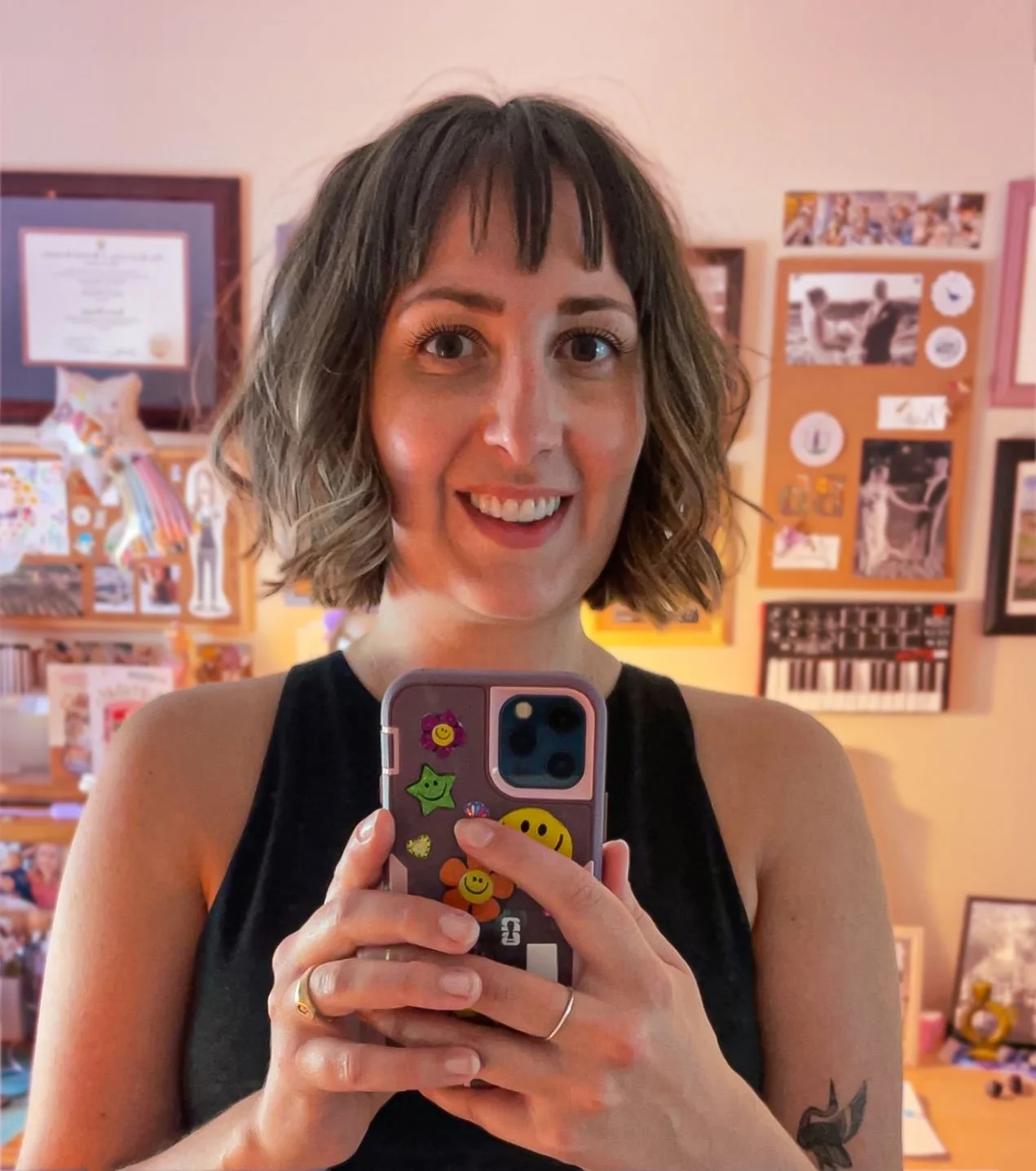 A woman with shoulder-length wavy hair and a black sleeveless top taking a selfie in front of a mirror. She is smiling and holding her phone decorated with colorful stickers. The background shows a room with framed pictures, posters, and a bulletin board.