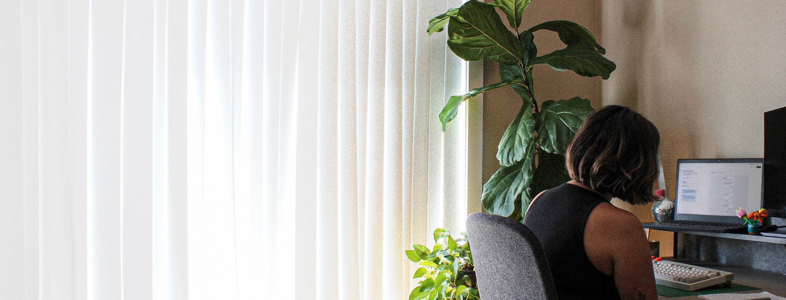 A woman working at a desk with a computer, large green plants, and white vertical blinds behind her.