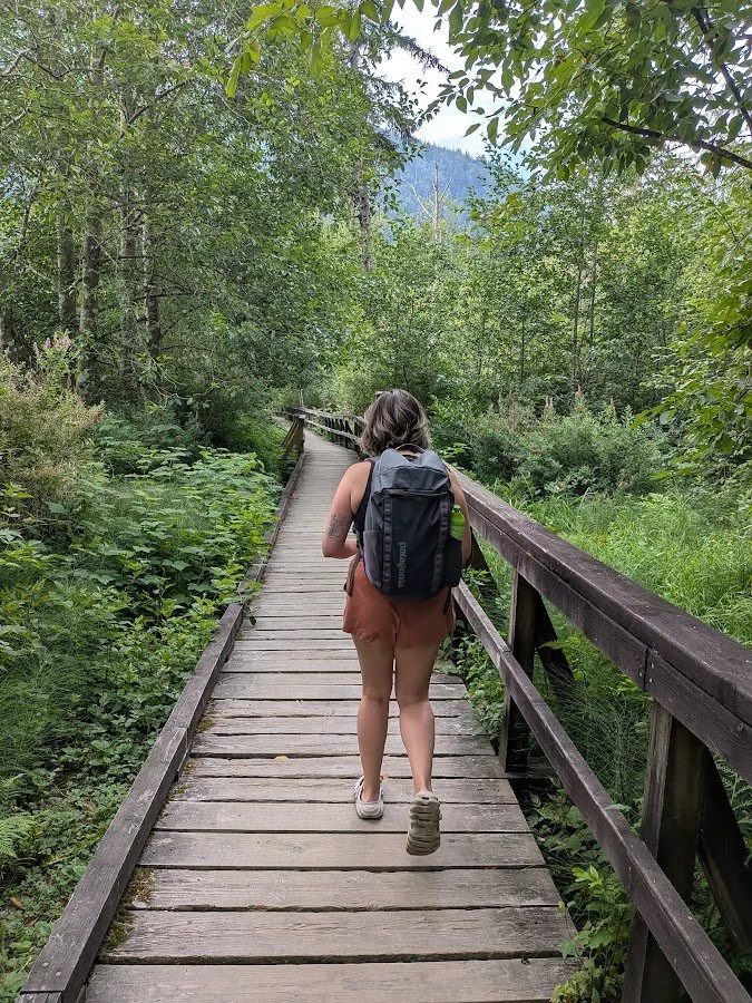 A woman walking on a narrow wooden trail through a lush green forest.