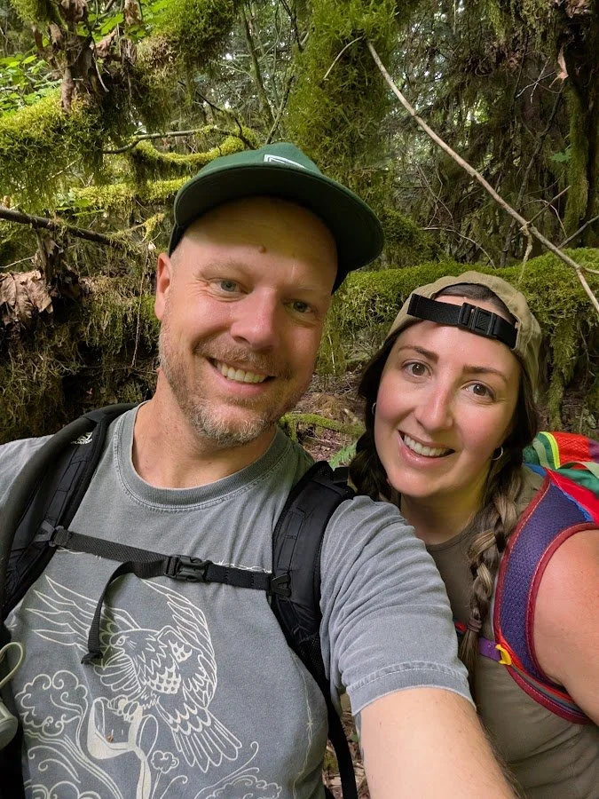 A man and woman smiling for a selfie in a forested area, surrounded by moss-covered trees and greenery. They are wearing casual outdoor clothing and backpacks, indicating they are hiking or exploring nature together.