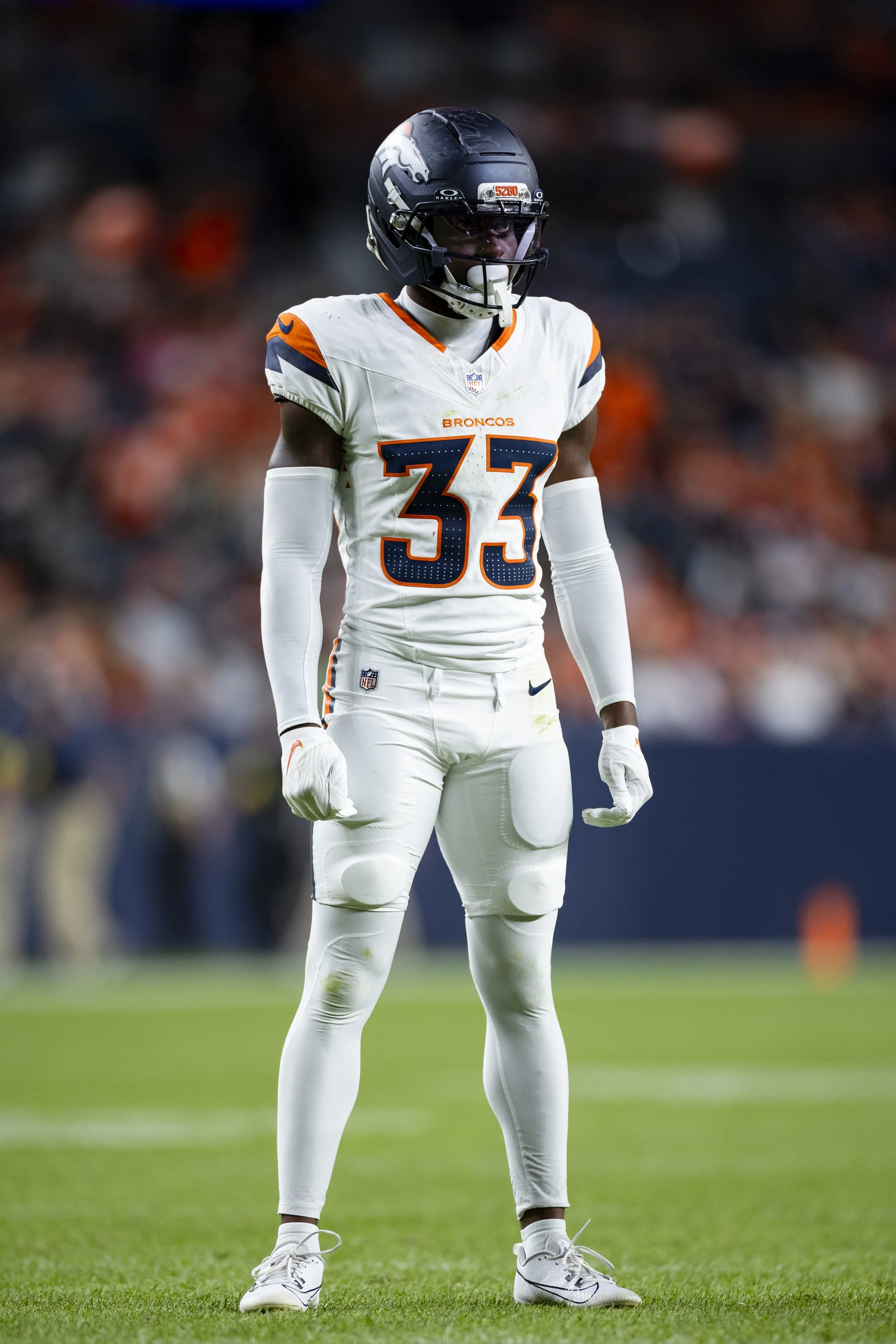 A football player in a Denver Broncos uniform standing on the field during a game, wearing a helmet and gloves.