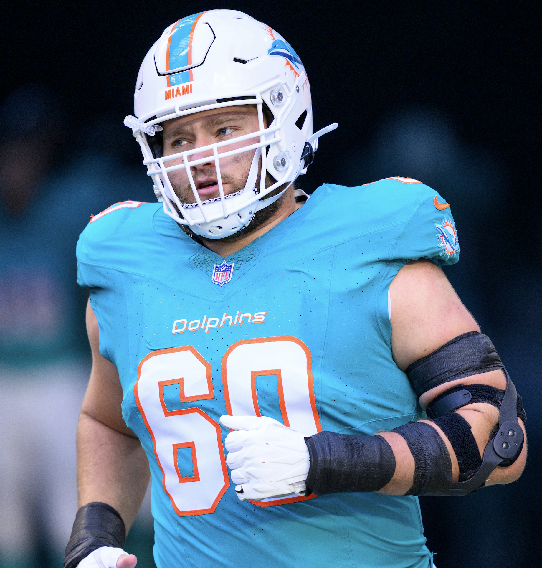 Close-up of a football player in a Miami Dolphins uniform with a white helmet, blue jersey, and orange accents, wearing a black arm sleeve and white gloves, on a football field.