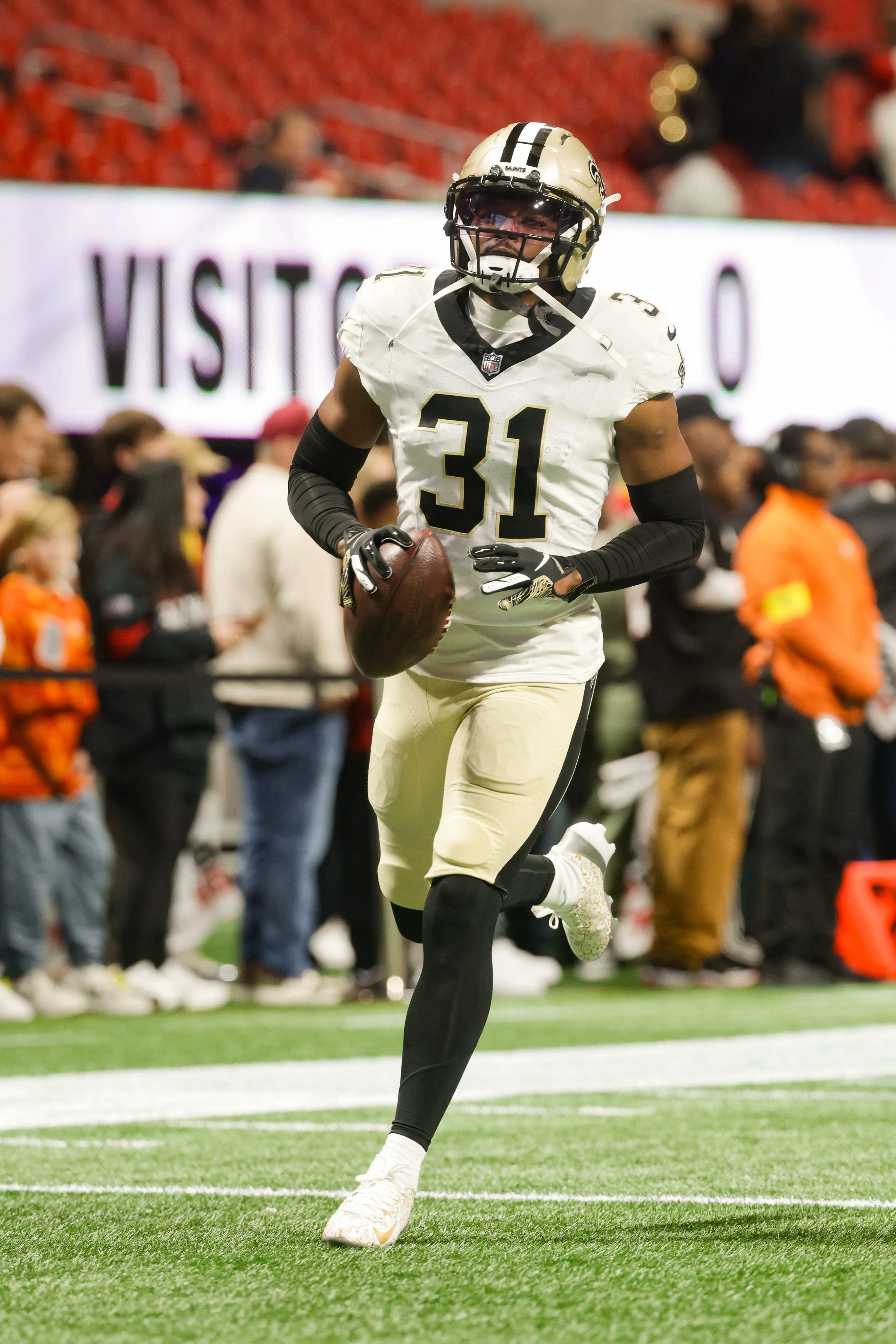A football player from the New Orleans Saints holding a football during a game, running on the field with spectators in the background.