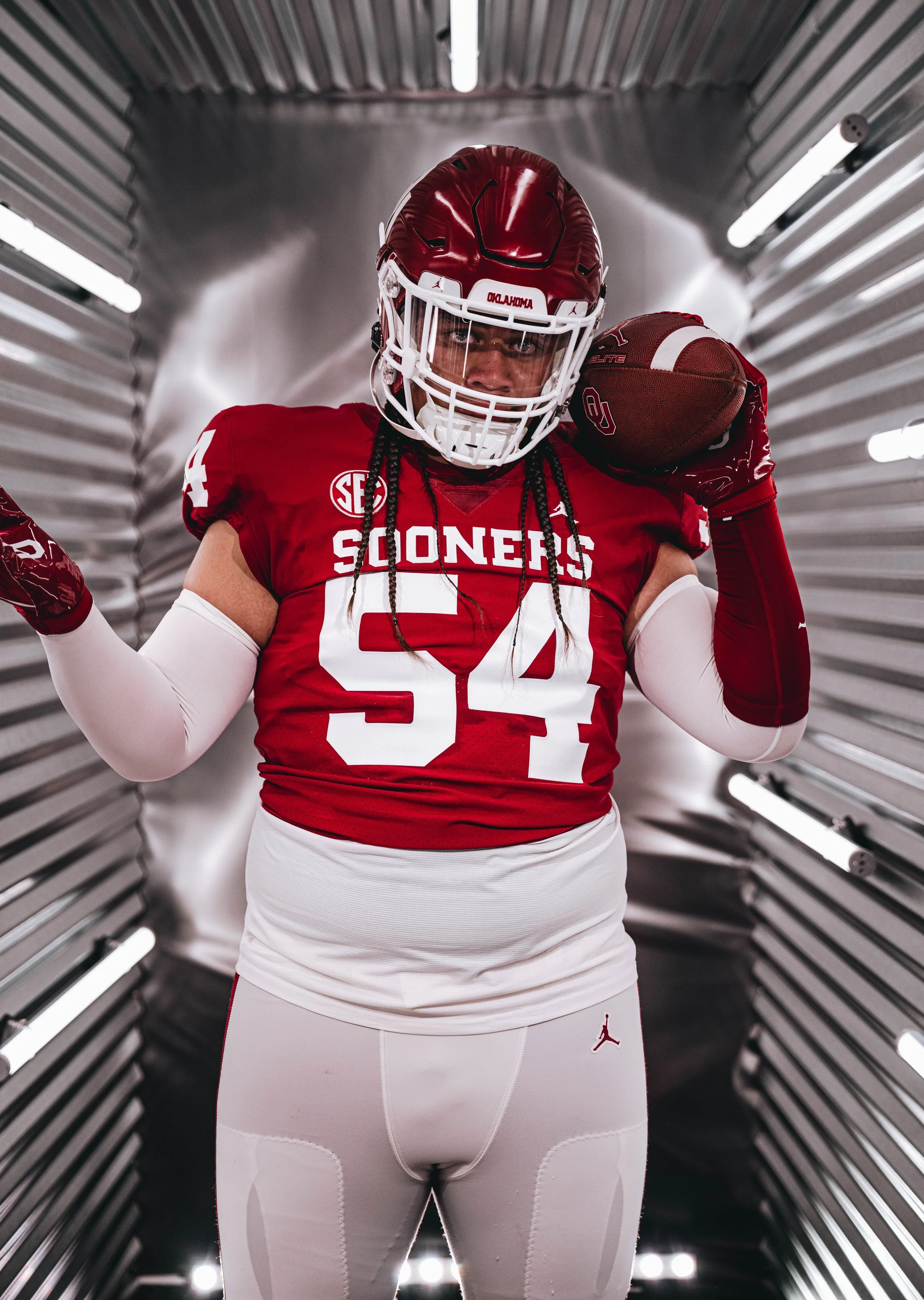A football player dressed in Oklahoma Sooners uniform holding a football inside a metallic tunnel, wearing a red helmet with a white faceguard.
