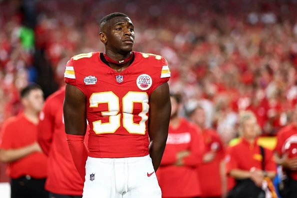 An African American football player wearing a red Kansas City Chiefs uniform with the number 30 stands on the field during a game, with a blurred crowd in the background.