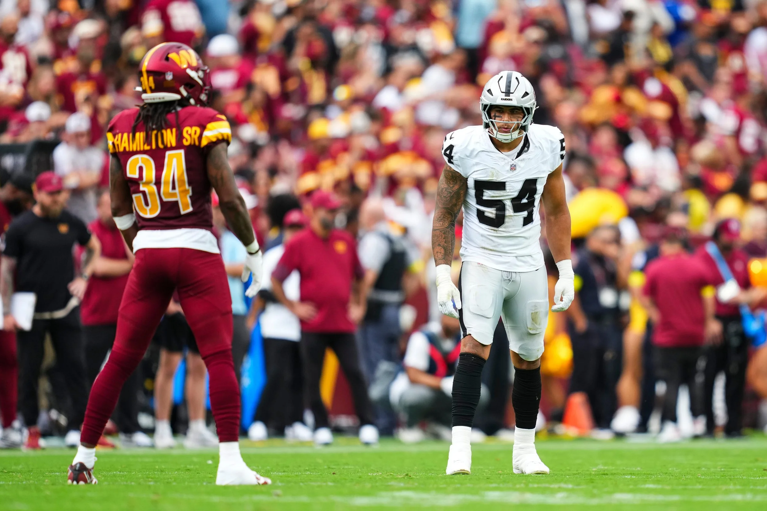 An American football game with two players on the field, one from the Washington Football Team and the other from the Las Vegas Raiders, standing and looking at each other, with a crowd of spectators in the background.