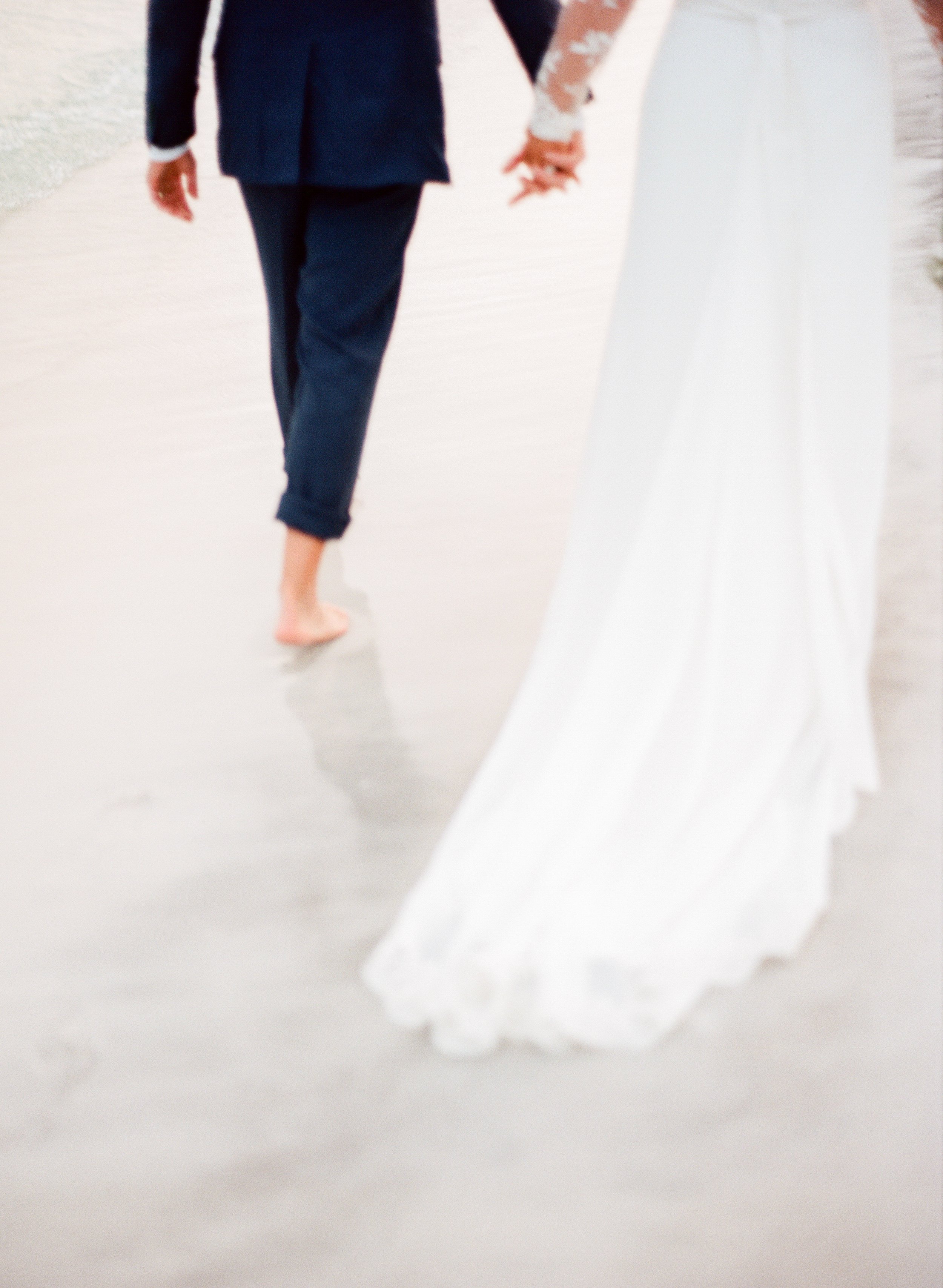 Beach destination wedding with bride and groom walking barefoot holding hands by luxury wedding photographer Amanda Watson