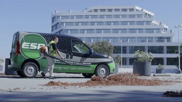 A person cleaning dirt off a black and green ESG service van parked on a city street with a modern white building in the background.
