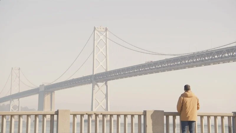 A man in a tan jacket standing by a railing, looking at a large suspension bridge over the water.