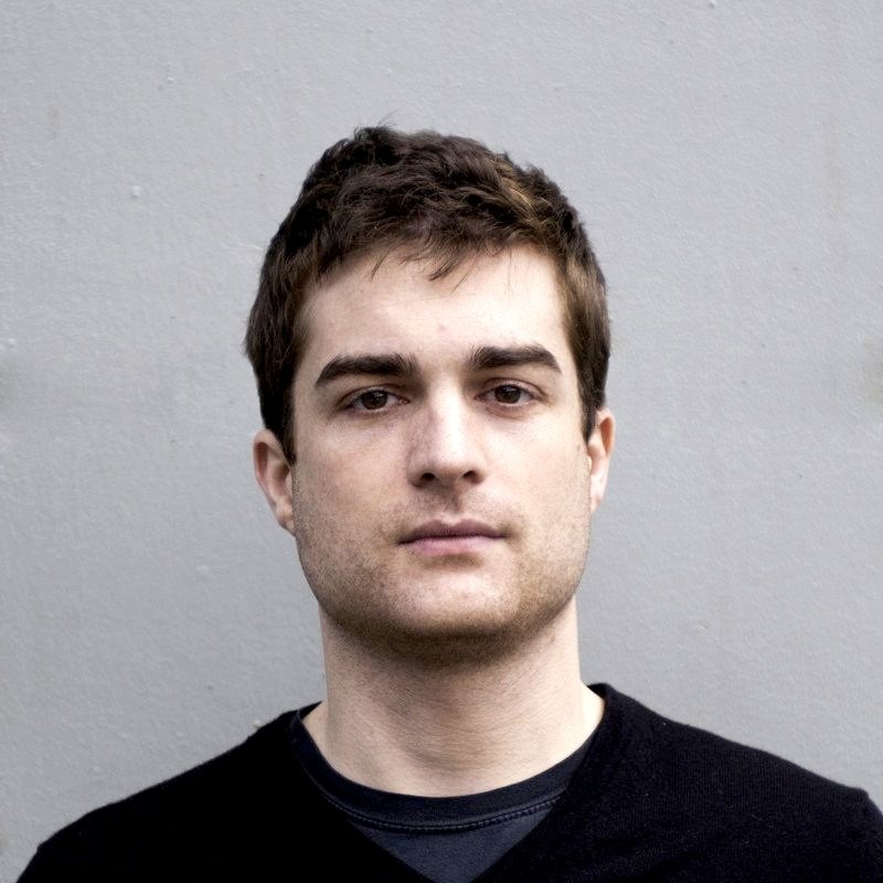 A portrait of a young man with short brown hair, wearing a black shirt, against a gray background.