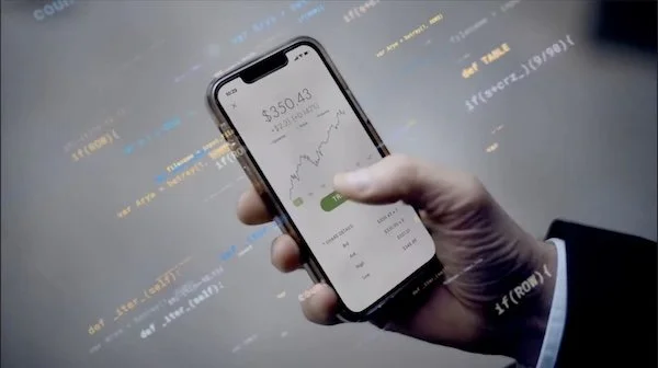 Close-up of a person's hand holding a smartphone displaying a financial app with a $350,043 balance and a rising graph, with computer code visible in the background.